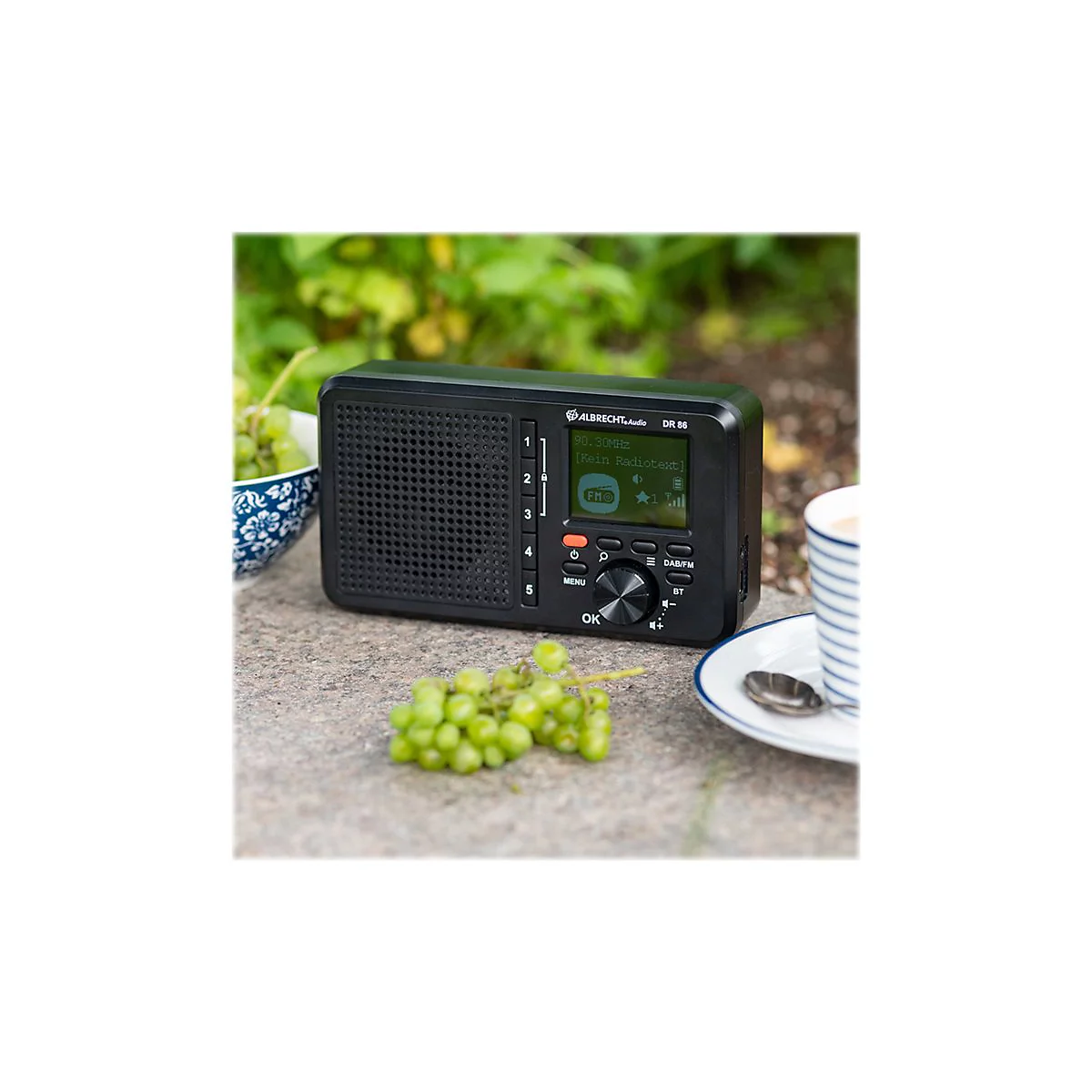 Schwarzes Radio mit Display, Trauben und Kaffee auf Steintisch. Hintergrund: GrÃ¼npflanzen.