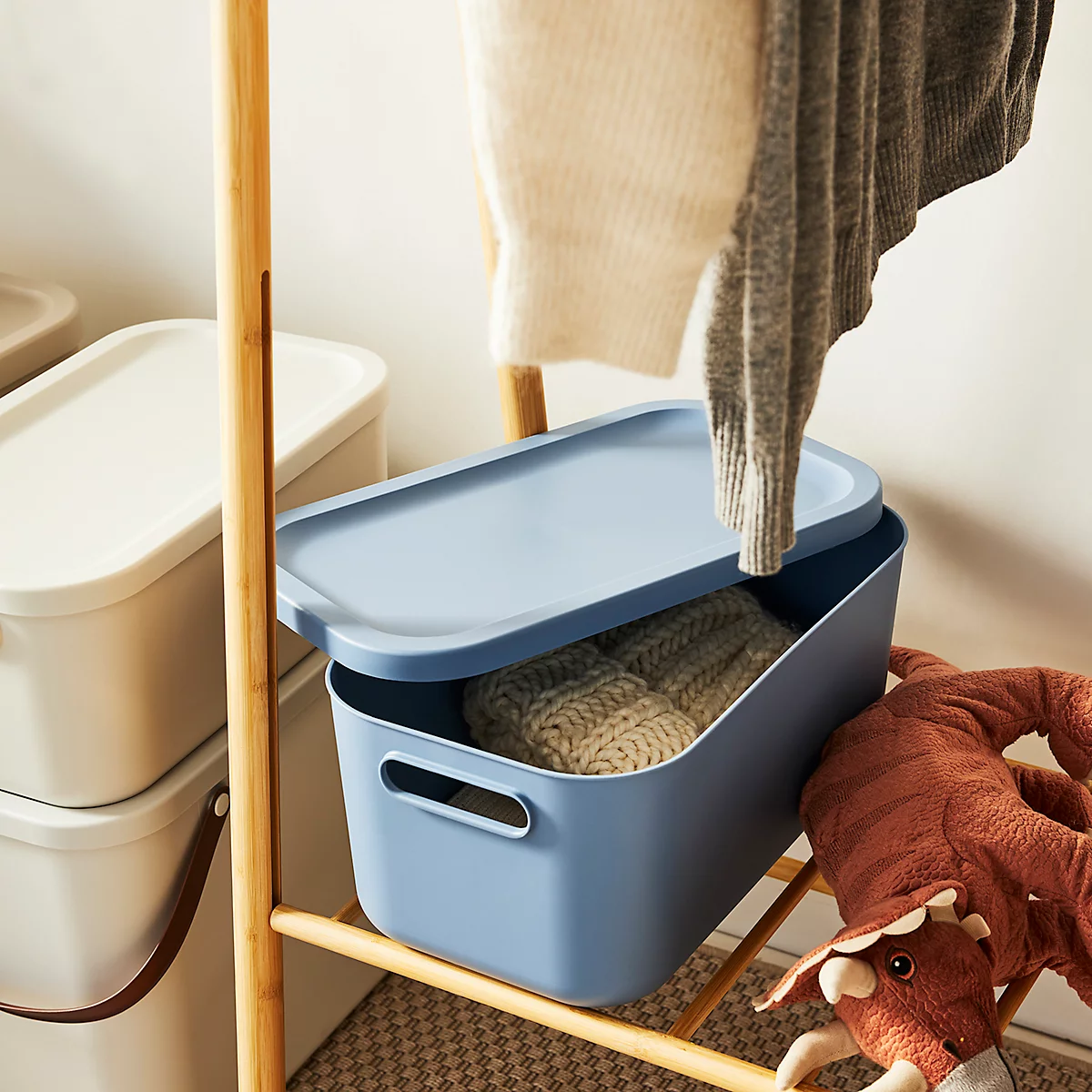 Blue storage box with lid, filled with knitwear, stands on a wooden rack. Clothing hangs above.