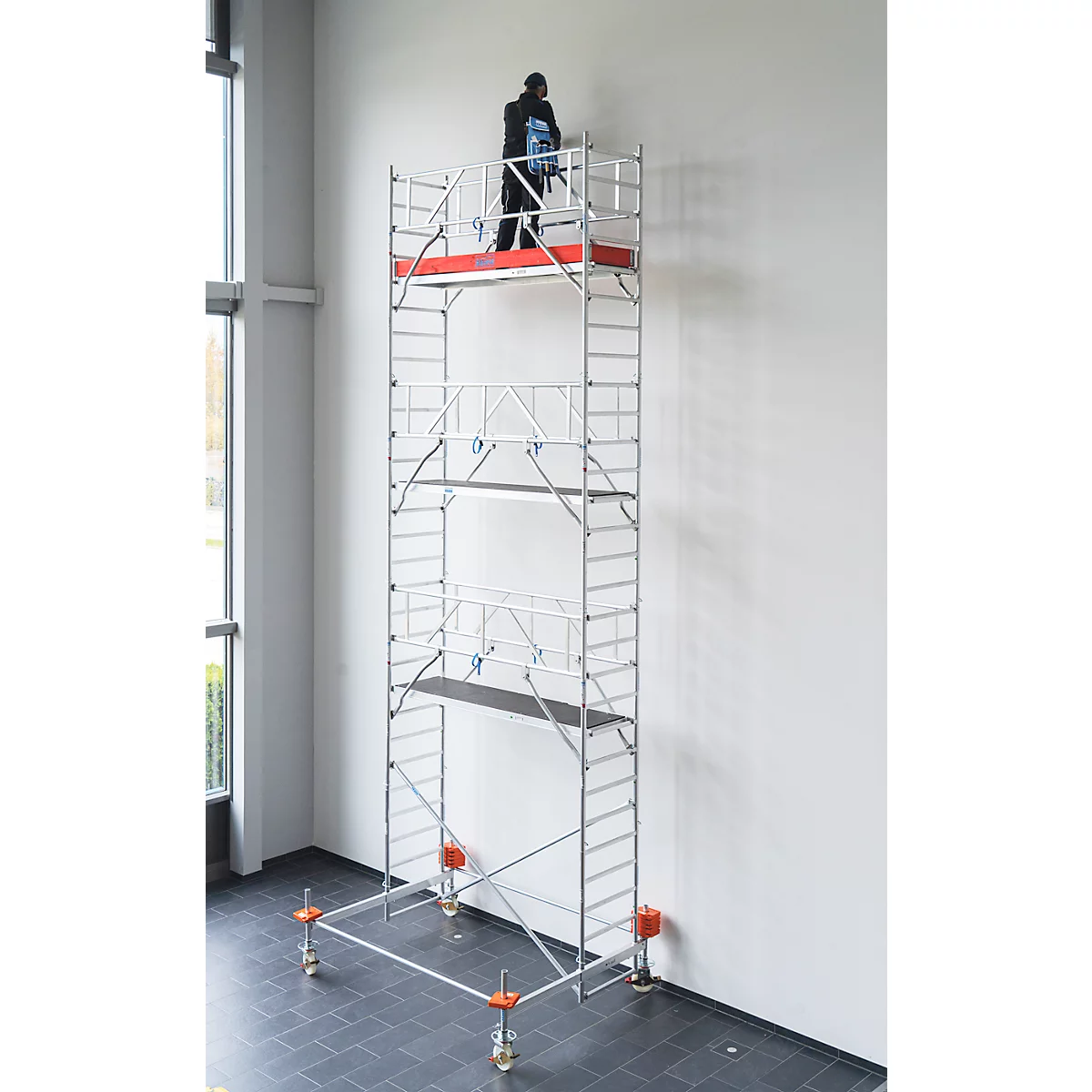 Person auf einem Gerüst arbeitet an einer weißen Wand. Graues Gerüst mit roten Akzenten und Rädern.