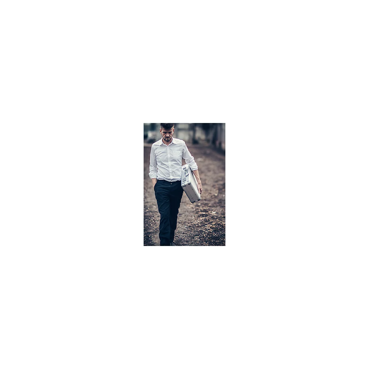 A man with a suitcase walks a path, serious look. He wears a shirt and pants.