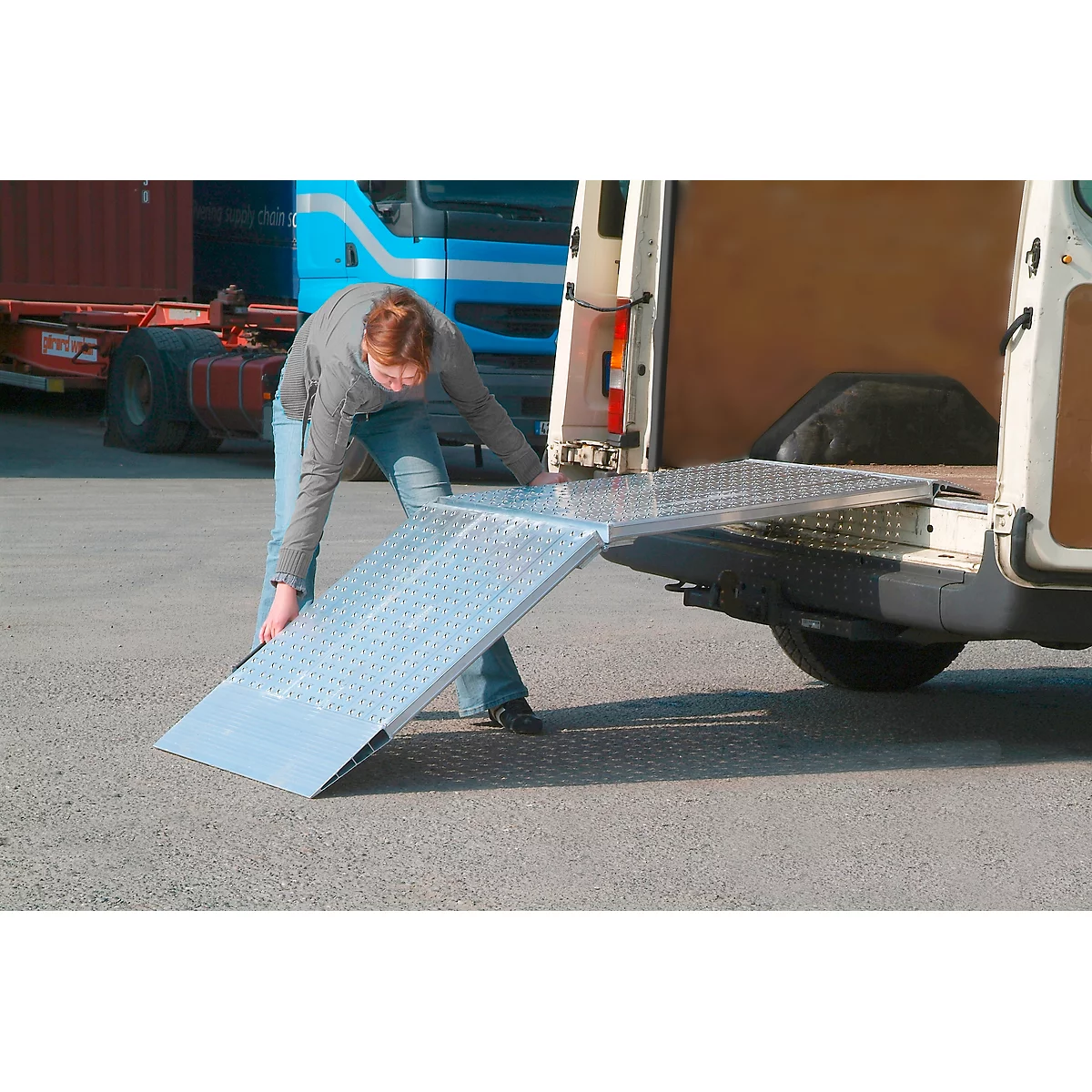 Person opening a loading ramp on a white van. A blue truck cabin can be seen in the background.