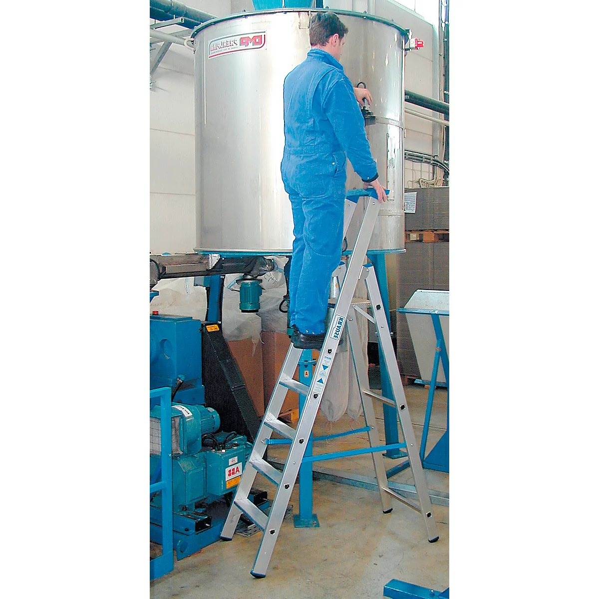 A man in blue overalls stands on a ladder in front of a large silver container.