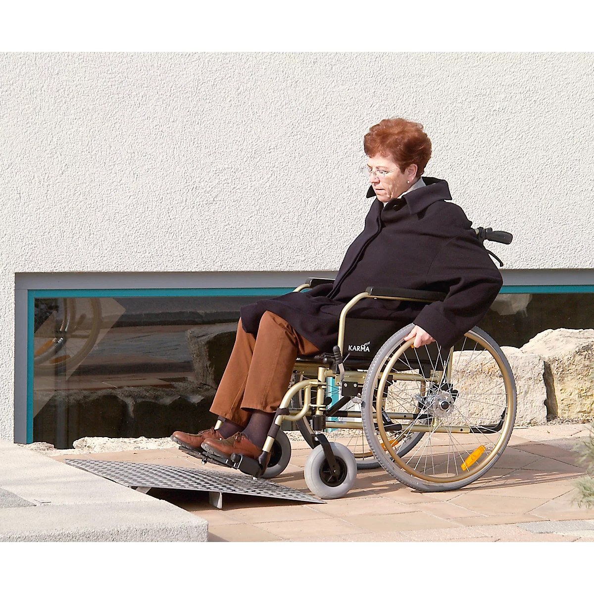 An elderly woman in a wheelchair drives over a ramp. She is wearing a black coat. In the background a window and stones.