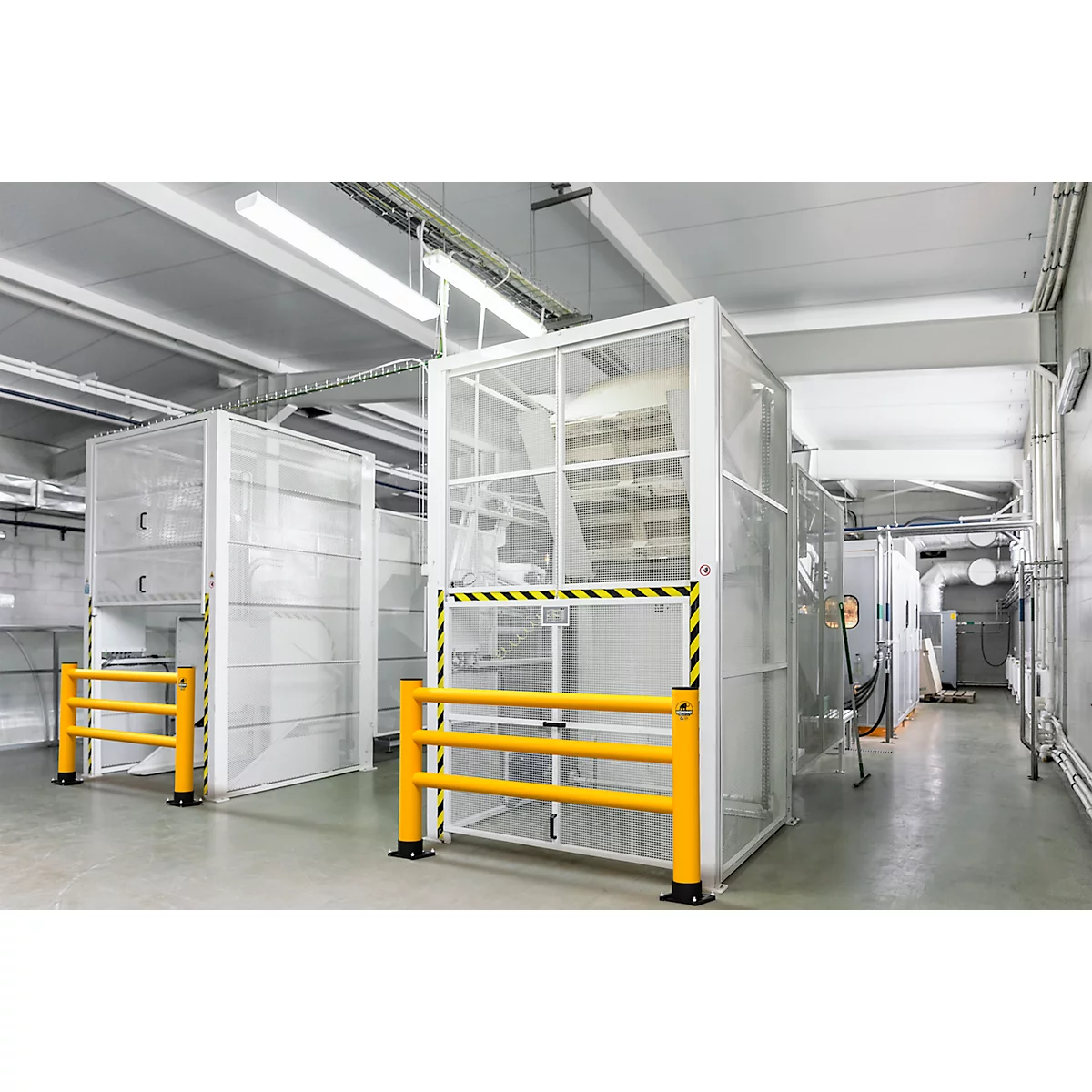 Industriehal met witte veiligheidskooien en gele afzettingen. Op de achtergrond buizen en ventilatie.