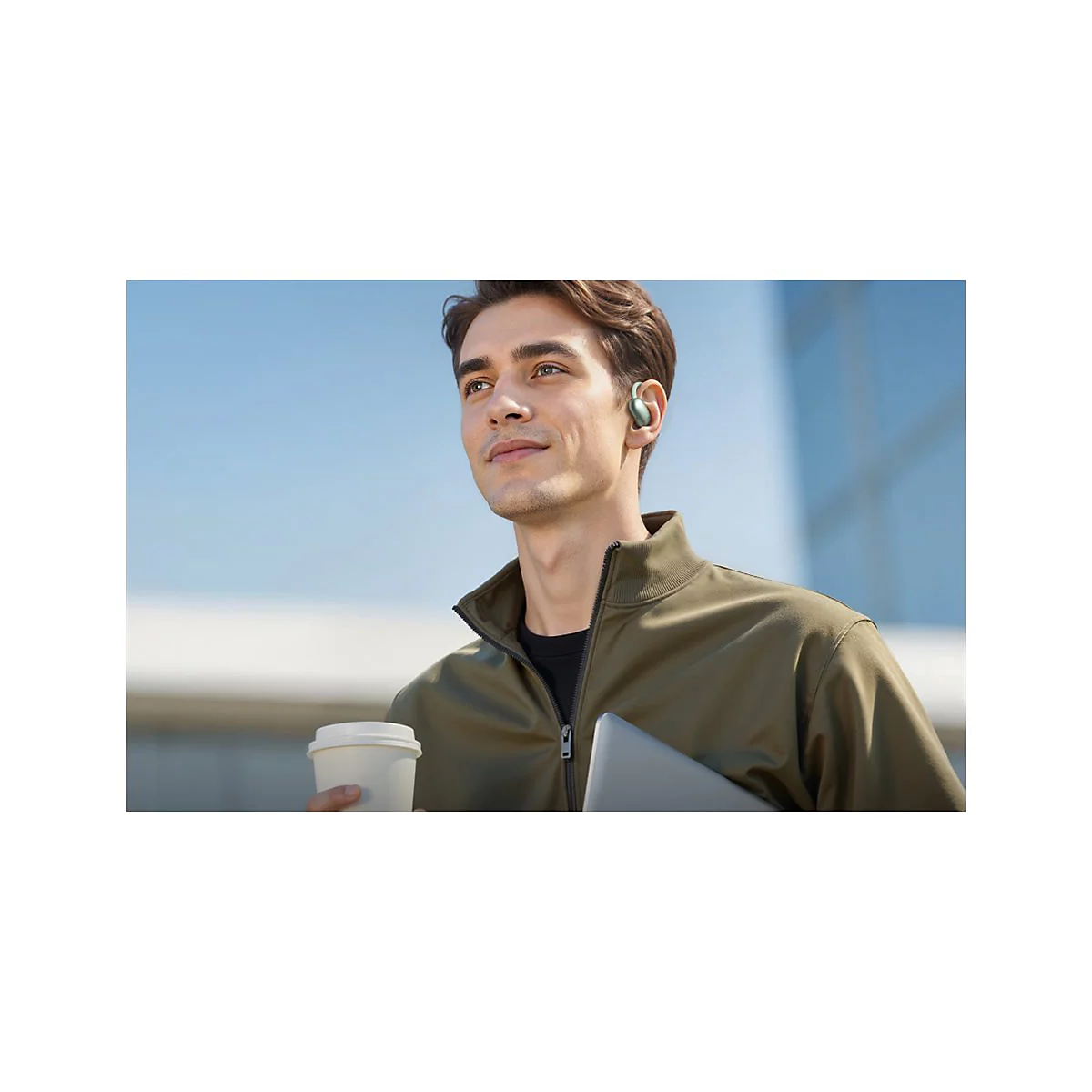 Ein junger Mann mit offenem Blick trägt einen grünen Ohrhörer und hält einen Kaffeebecher in der Hand.
