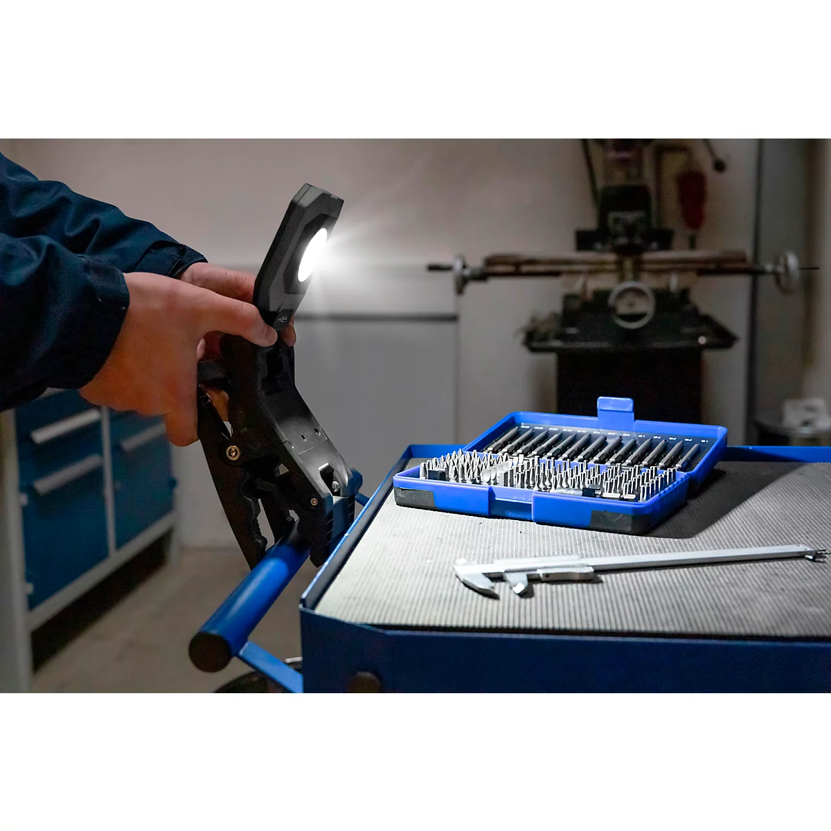 Een monteur die een zwarte werklamp met helder licht in de werkplaats vasthoudt. Op de achtergrond gereedschap, blauwe doos met opzetstukken.