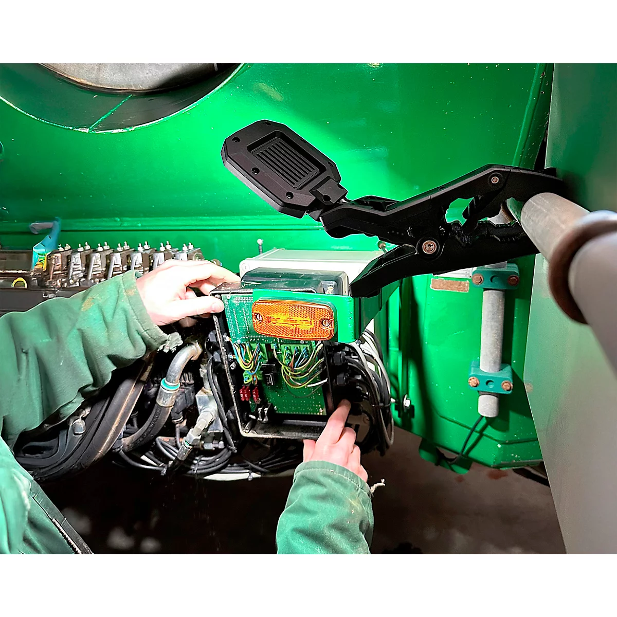 Een persoon in groene werkkleding repareert elektronica onder een lamp met klem. Een oranje knipperlicht is zichtbaar.