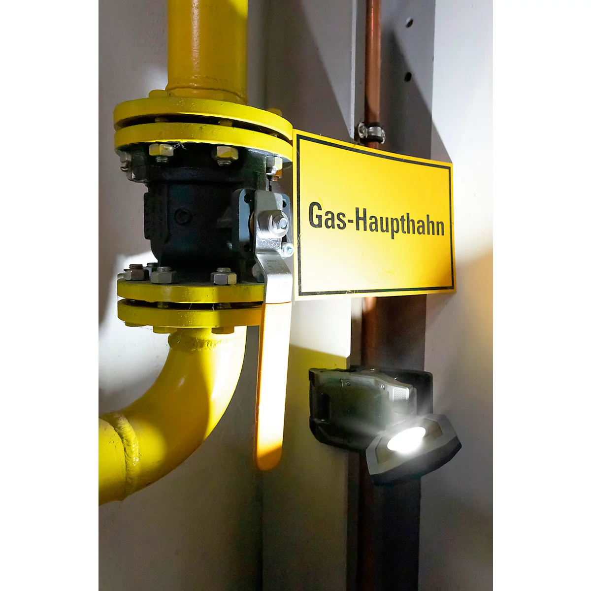 Gele gasleiding met afsluiter en bordje "Gas-Haupthahn". Een licht schijnt op de scène.