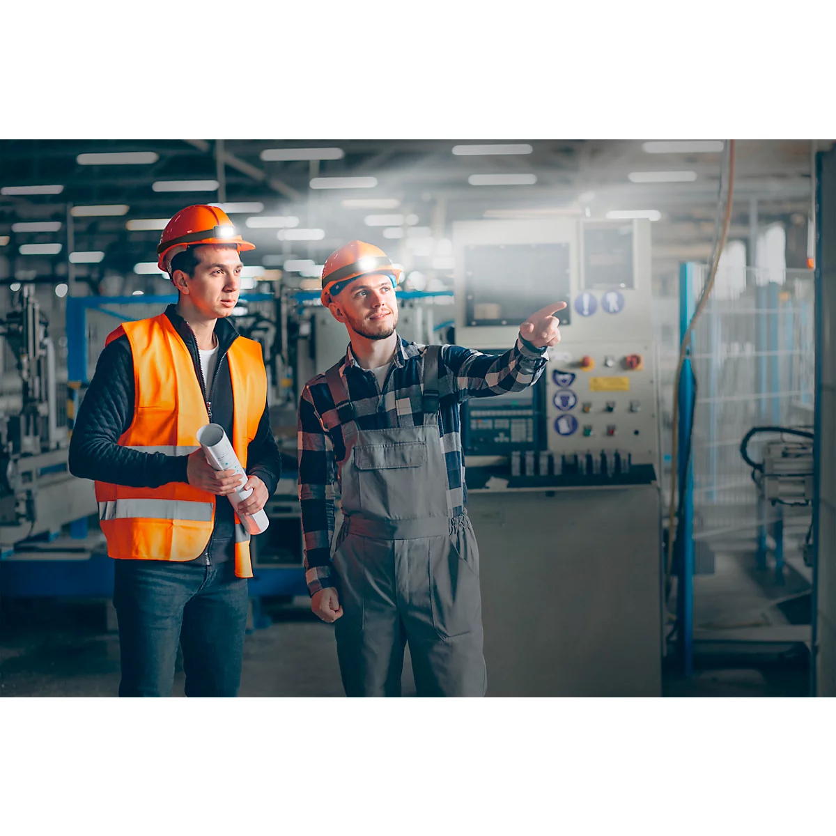 Two men in helmets and work clothes in a factory. One is pointing at a machine, the other is holding plans.