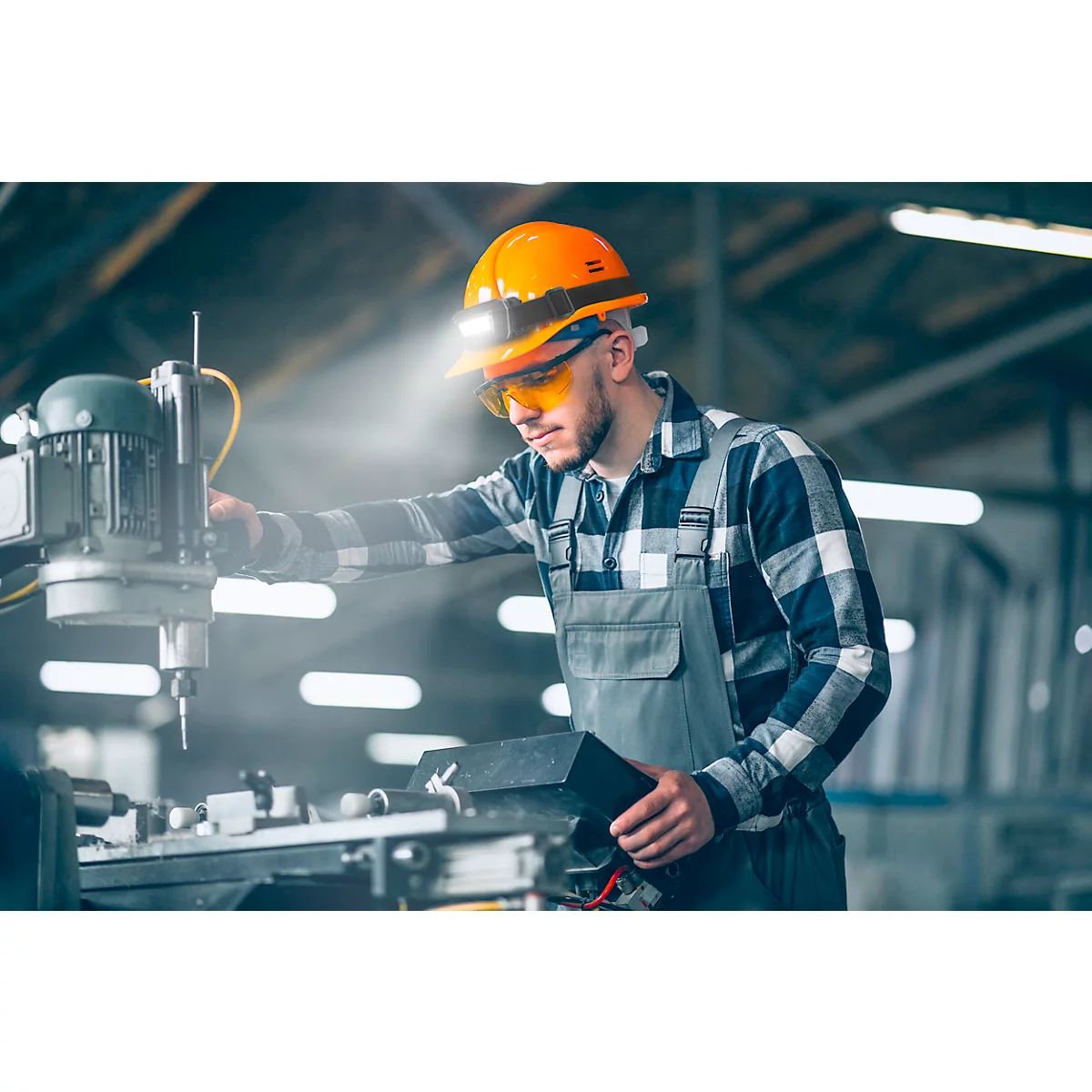 A worker in an orange helmet with LED light and safety glasses at a machine. He is holding a black device in his hand.