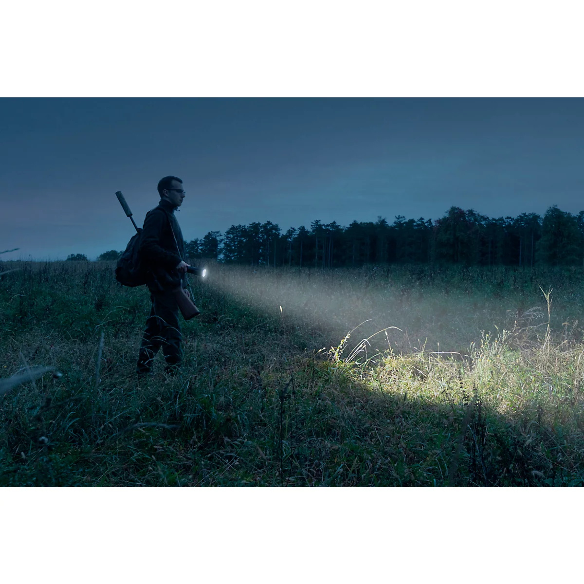 Person with rifle and backpack in field at night, flashlight illuminating the way.
