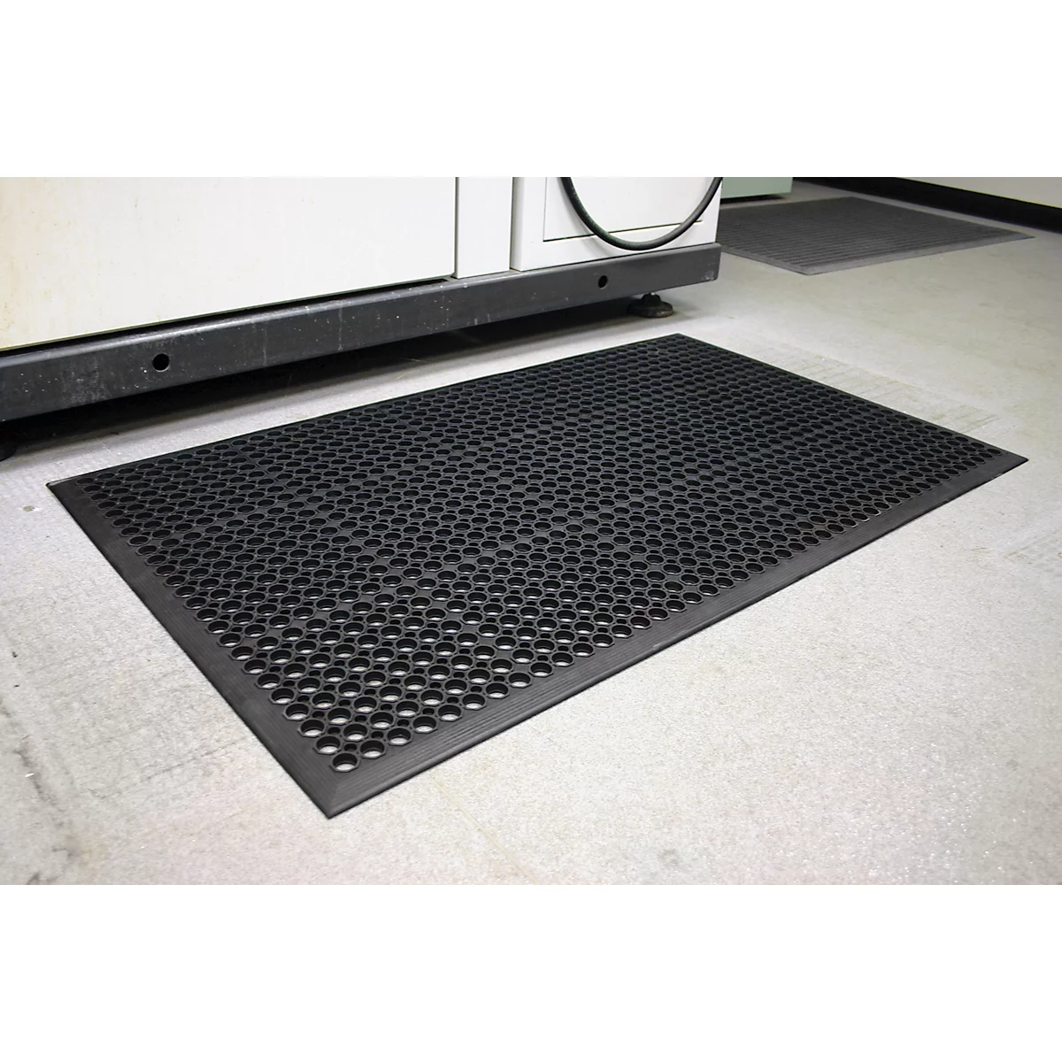 Black rubber mat with circular holes lies in front of a machine on a concrete floor.