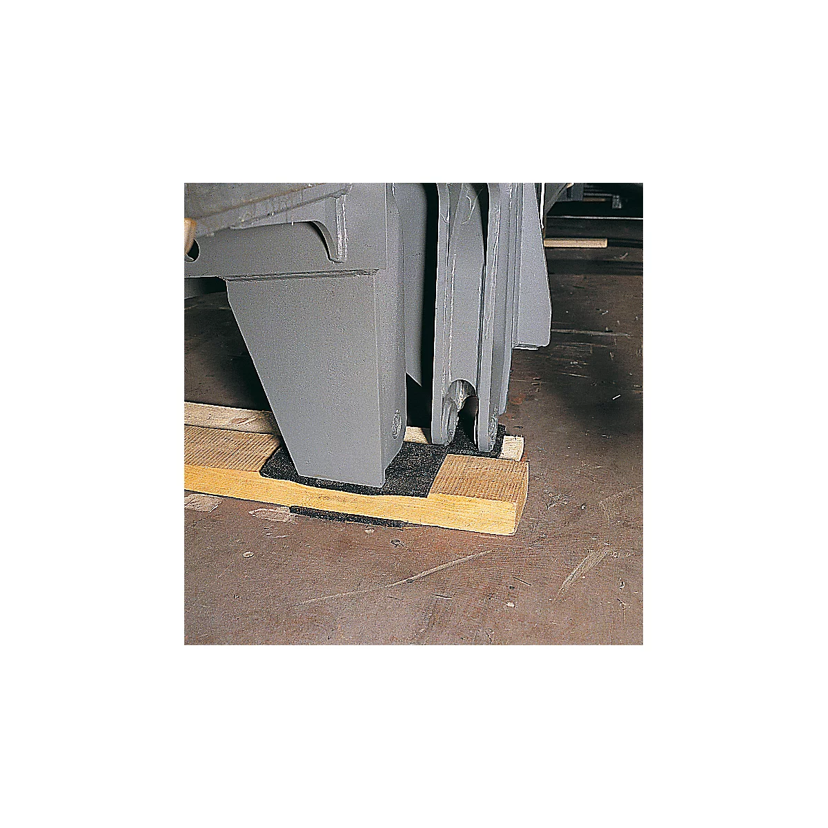 Gray machine part stands on a wooden block with a black base. Brown floor.