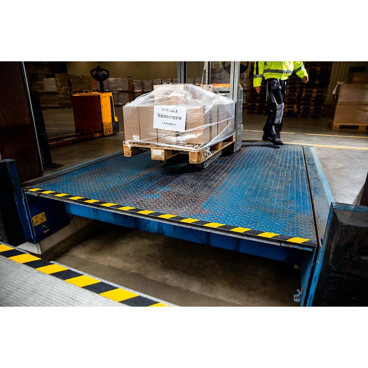 A worker in work clothes stands next to a loading ramp on which a package stands on a pallet.