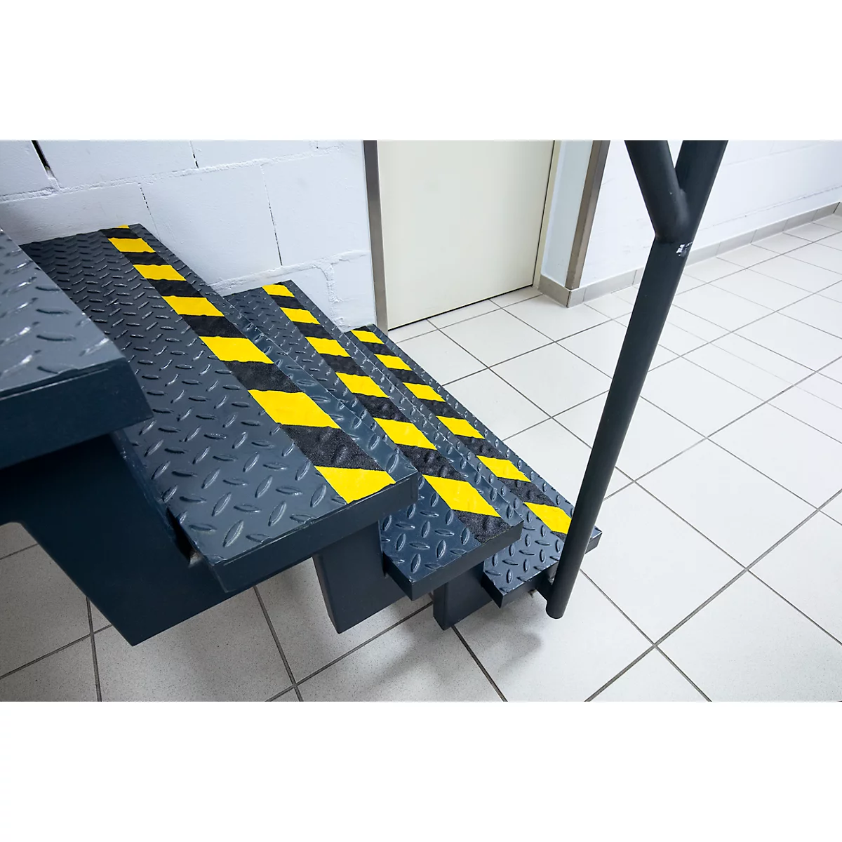 Steel staircase with yellow and black warning stripes. Handrail on the right, door and tiles in the background.