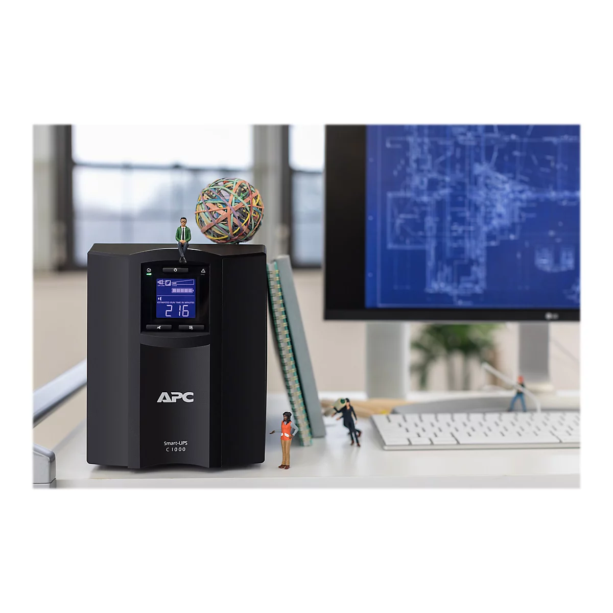 Ein schwarzes APC Smart-UPS C 1000, auf dem ein kleiner Mann sitzt. Auf dem Bildschirm steht "216" für die verbleibende Laufzeit. Auf dem Tisch sind auch kleine Figuren und Büromaterialien zu sehen.