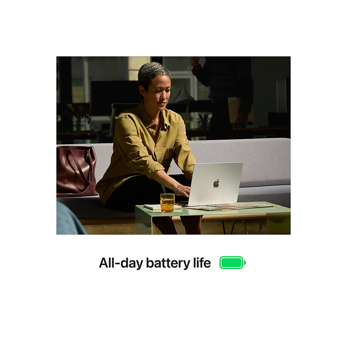 Frau sitzt an Tisch mit Laptop, im Hintergrund weitere Person. Auf dem Bildschirm ist das Apple-Logo zu sehen. Auf dem Tisch steht ein Glas.