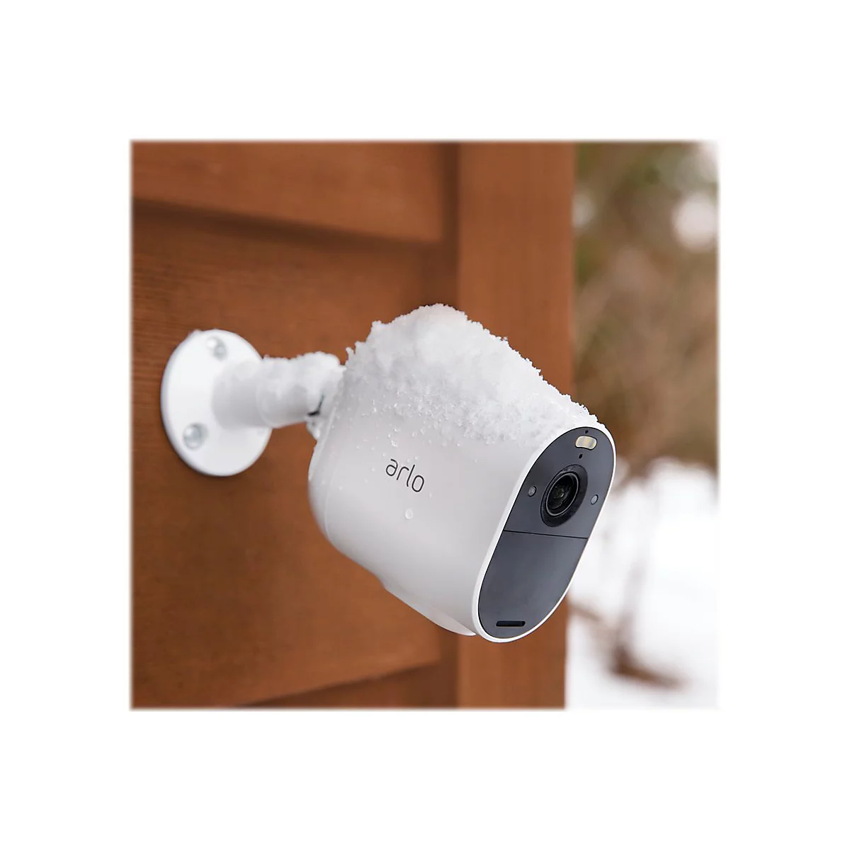 Eine weiße Arlo Überwachungskamera, bedeckt mit Schnee, ist an einer braunen Holzwand montiert.