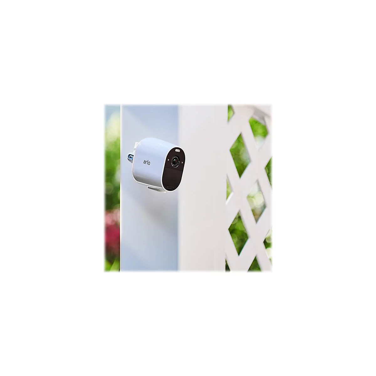 Eine weiße Arlo-Überwachungskamera, an einer Wand befestigt. Der Hintergrund zeigt einen weißen Gitterzaun mit unscharfem Grün.