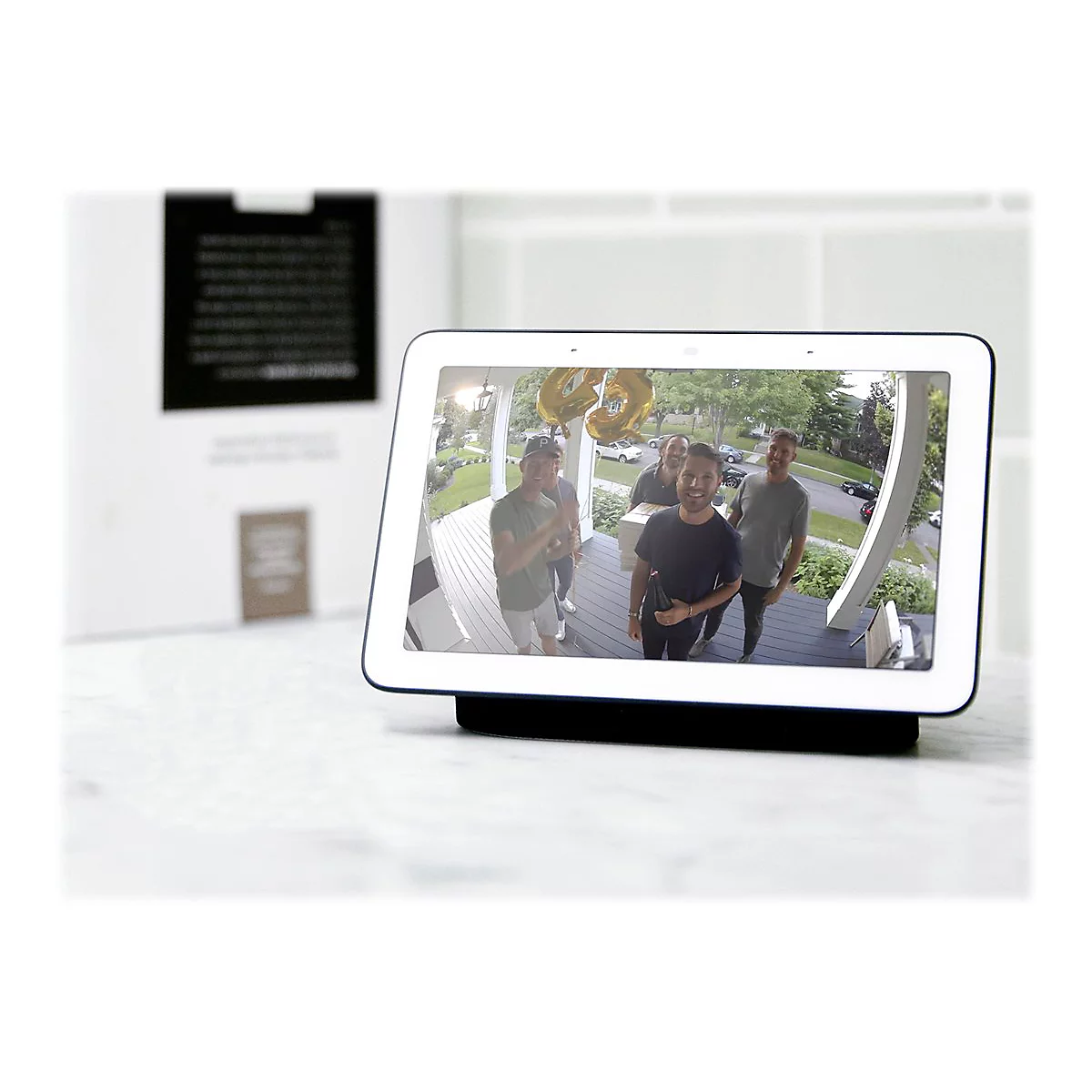 Ein Smart Display zeigt eine Aufnahme von Freunden, die vor einer Haustür stehen und lächeln. Goldene Luftballons. Das Display steht auf einem Tisch.