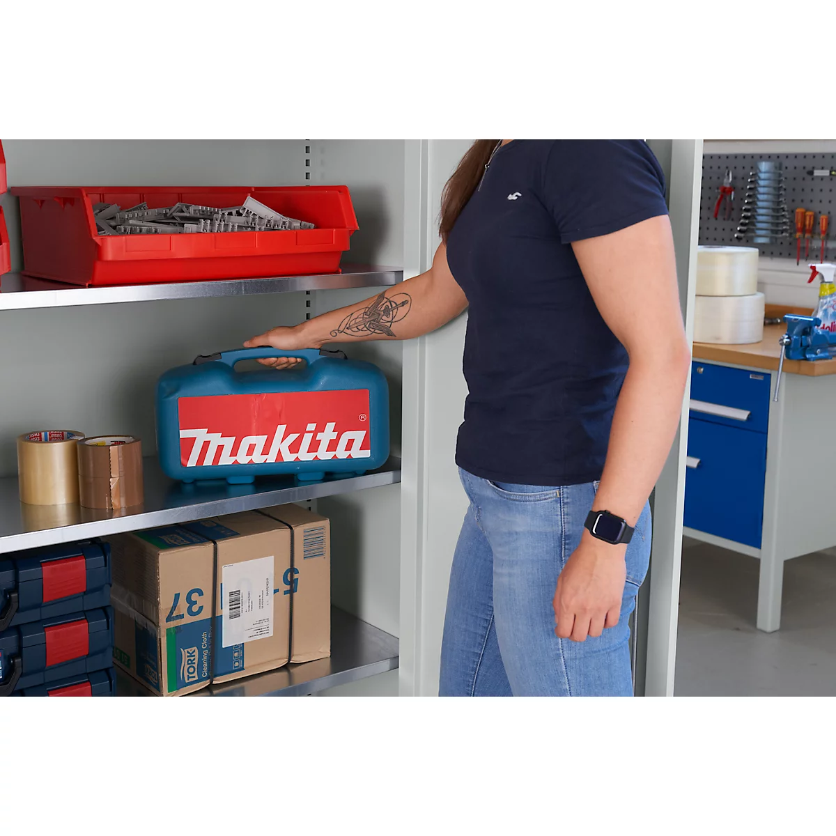 Una mujer en jeans y camiseta azul está de pie junto a un armario con una caja de herramientas. El logotipo de Makita es claramente visible.