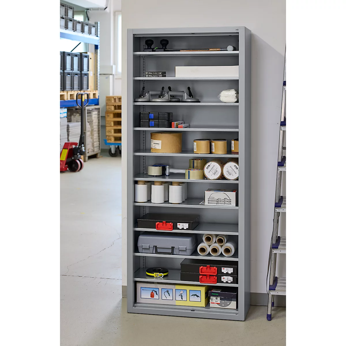 Armoire à outils grise avec des outils et du matériel dans un atelier. Derrière, un chariot élévateur et une échelle.