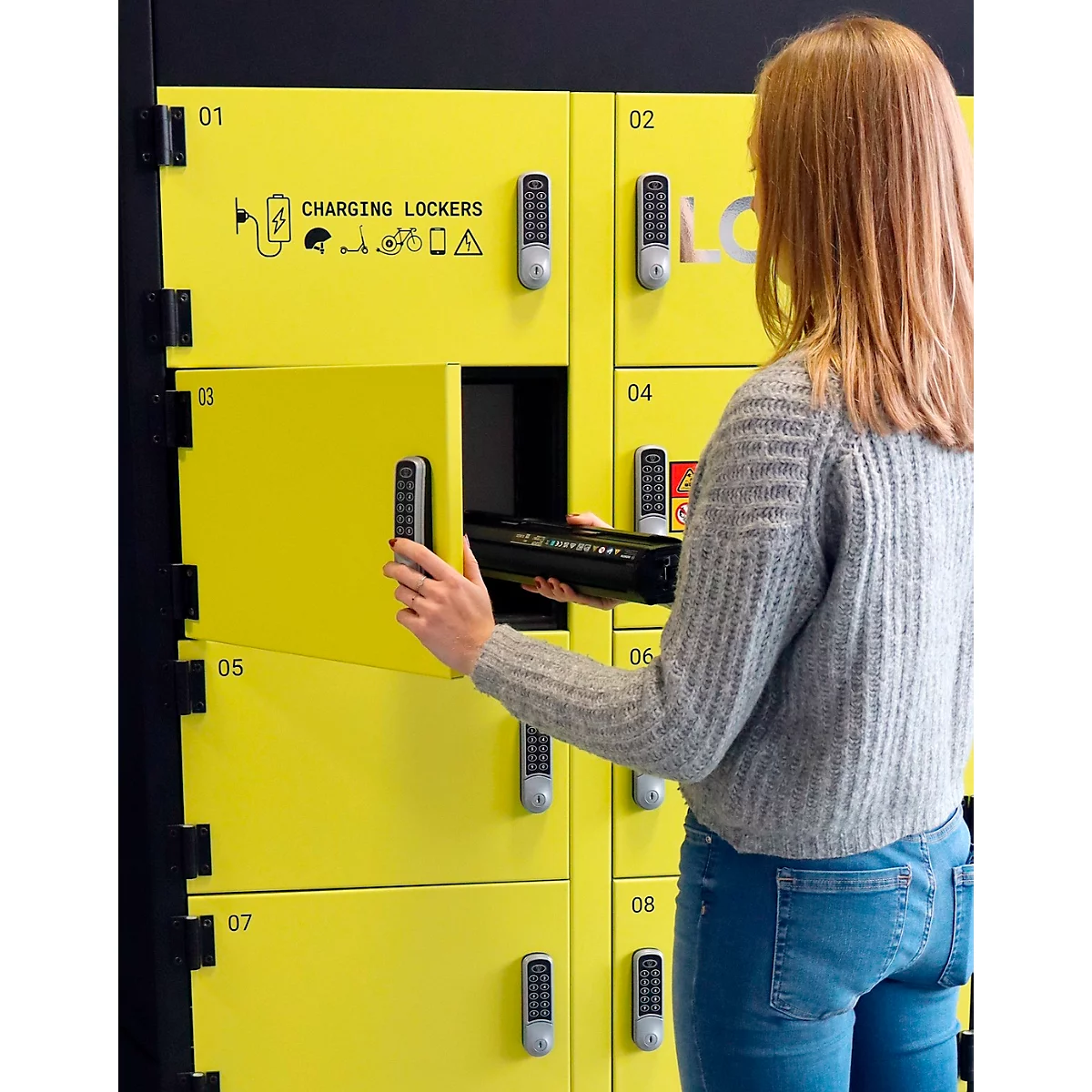 Jeune femme ouvrant un casier jaune. Sur le casier, il est écrit "CHARGING LOCKERS". Elle tient un objet noir.