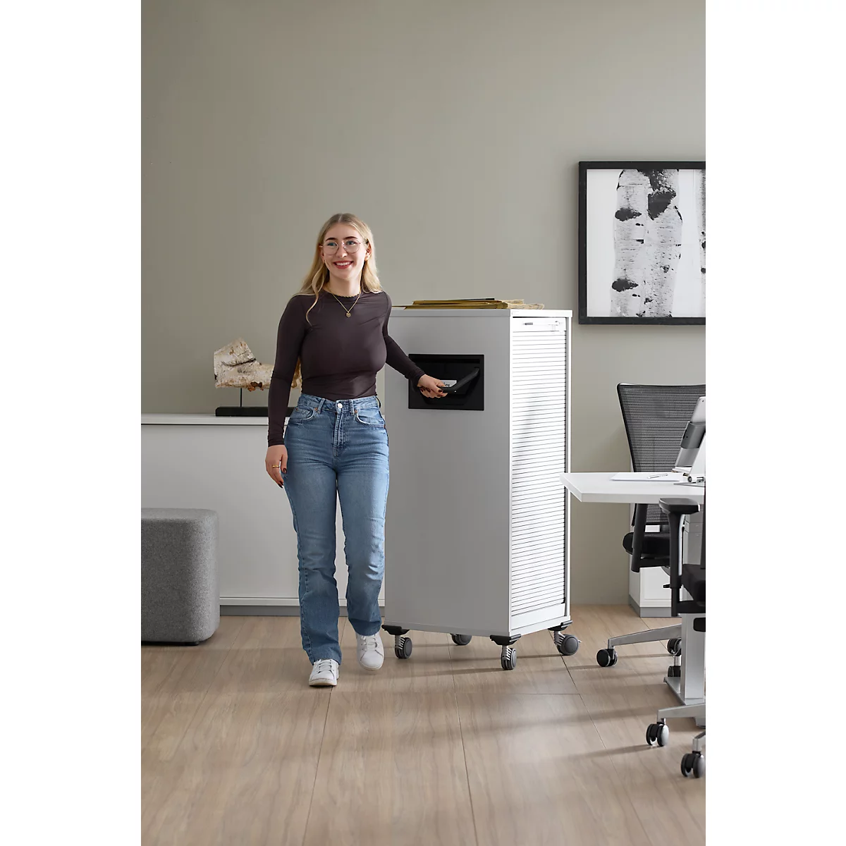 Une femme souriante se tient devant une déchiqueteuse de bureau. Elle tient un dossier.