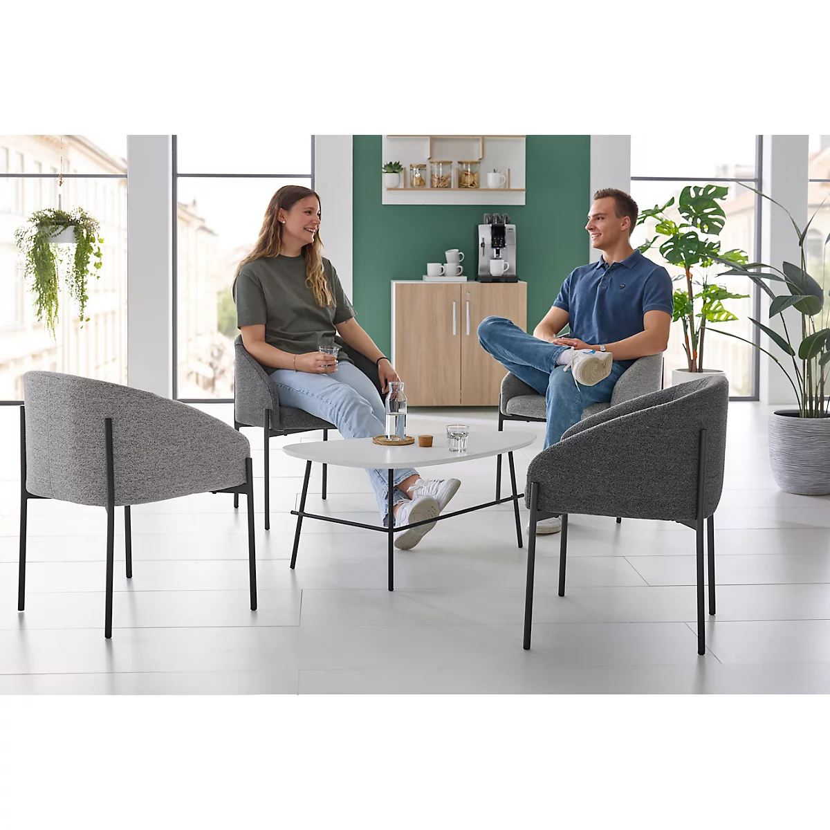 Two people sit in armchairs at a table. In the background a coffee machine and a cabinet.