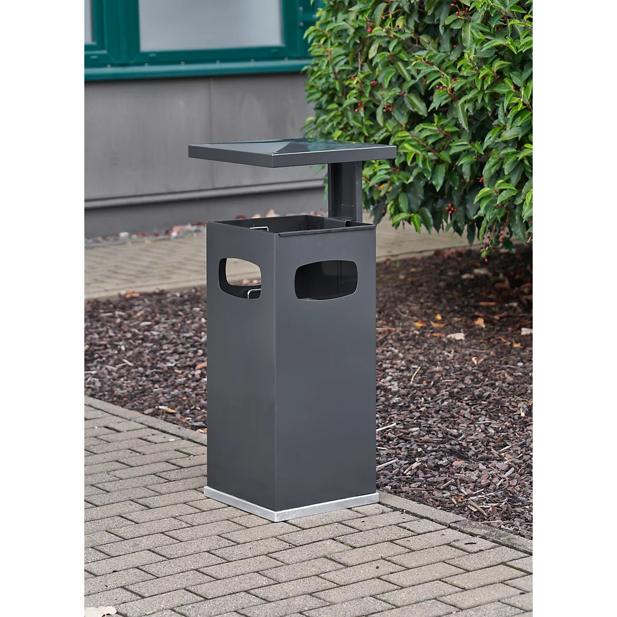 Grey outdoor trash bin with shelf, rectangular with cutouts. Standing on paving stones next to green areas.