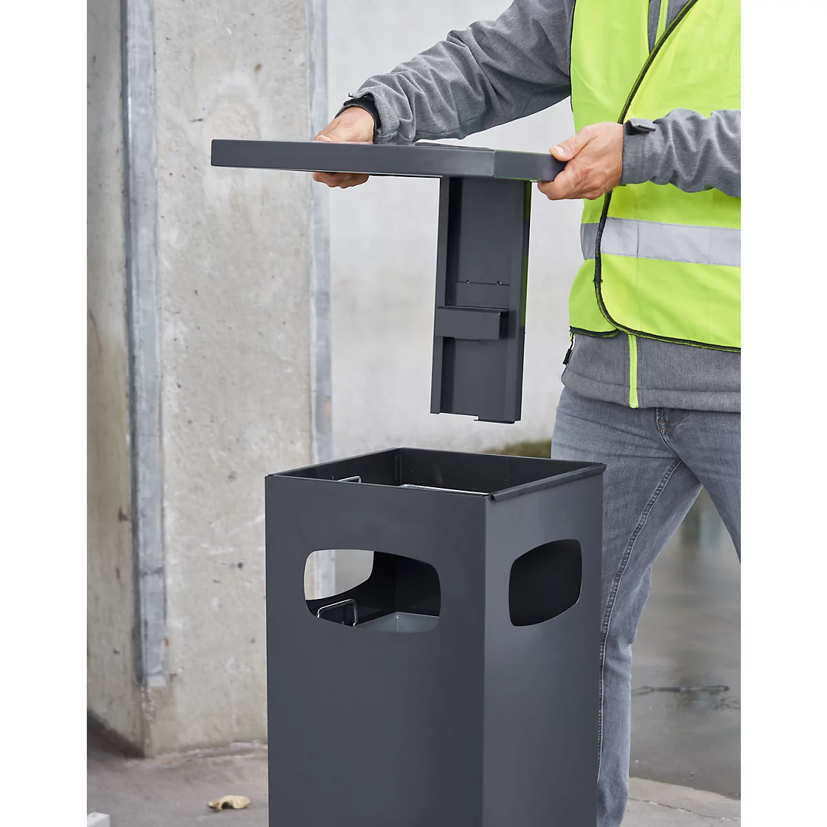 Person assembling grey waste container. The lid is being inserted. The man is wearing a yellow vest.