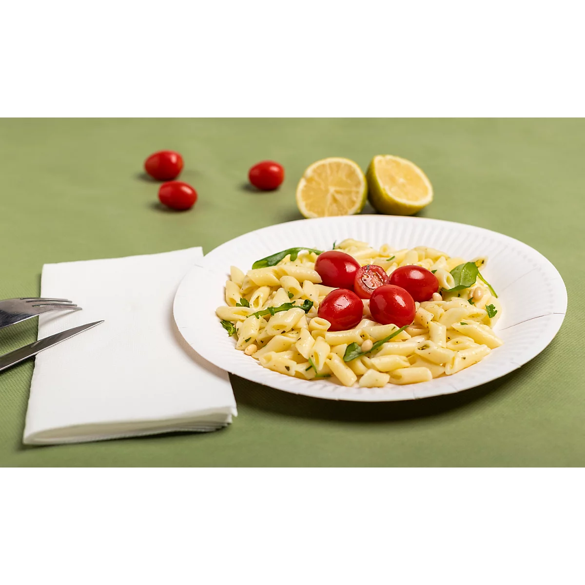 Pâtes avec tomates cerises et basilic sur une assiette en papier blanche, avec des moitiés de citron et des couverts sur une surface verte.