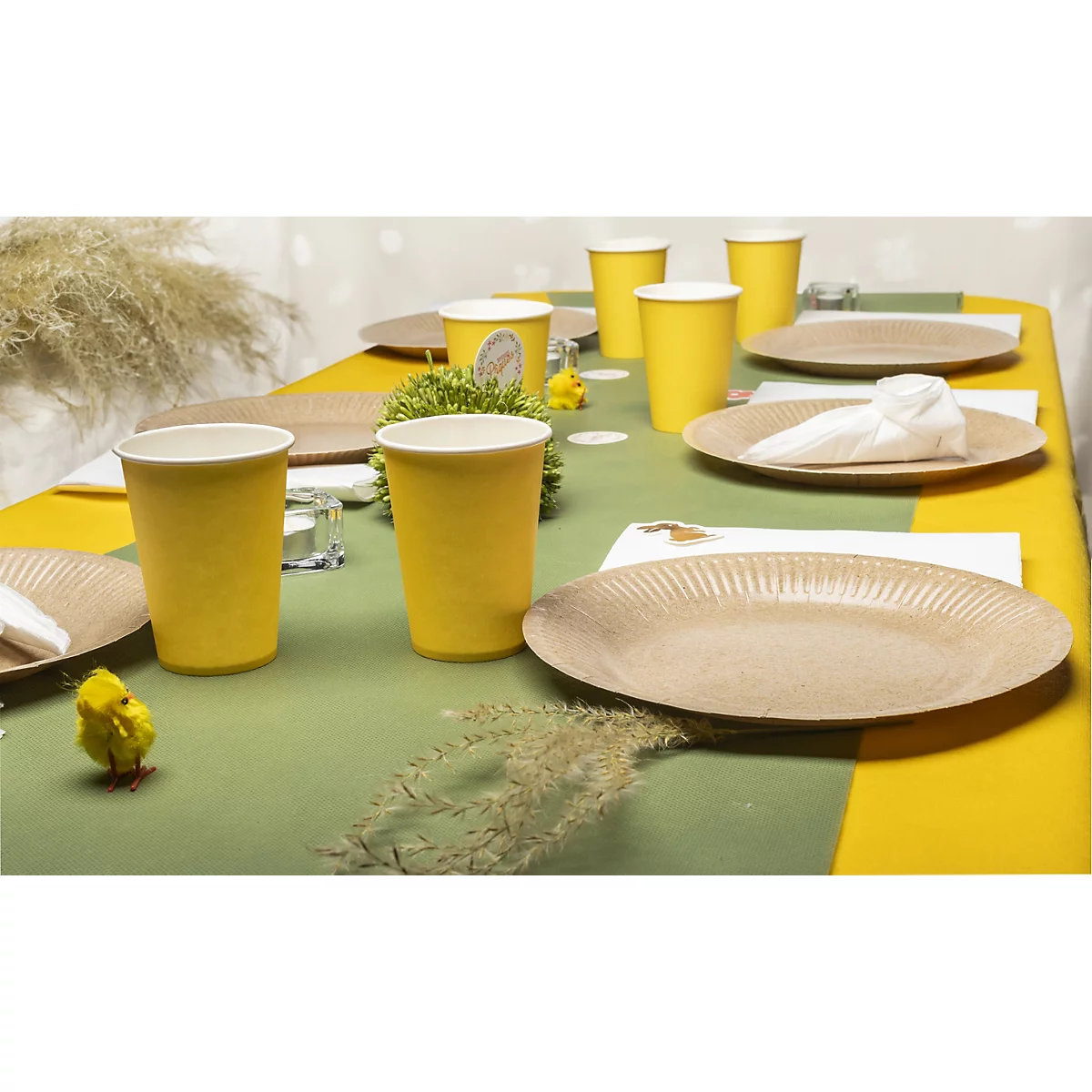 Table festive avec des gobelets, des assiettes et des serviettes jaunes. Décoration de Pâques avec des poussins et de la verdure.