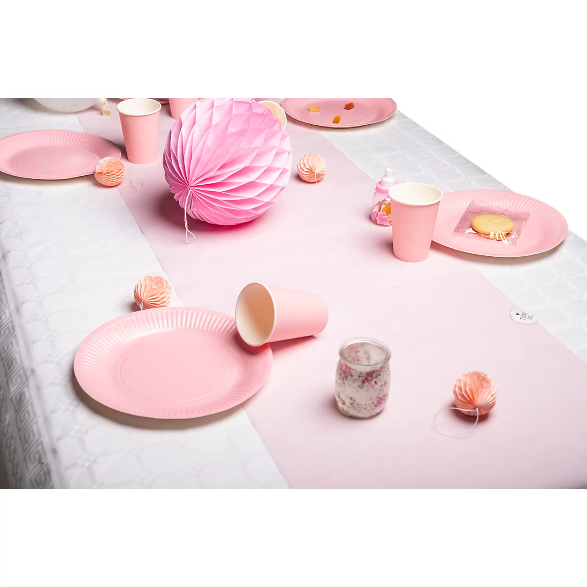 Une table dressée de manière festive avec des assiettes, des tasses et des décorations roses. Un chemin de table rose et une grande boule en papier sont sur la table.