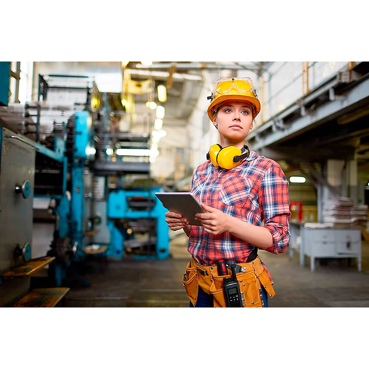 Jeune femme dans un atelier avec une tablette et un casque de sécurité. Regardant au loin, prête à travailler.