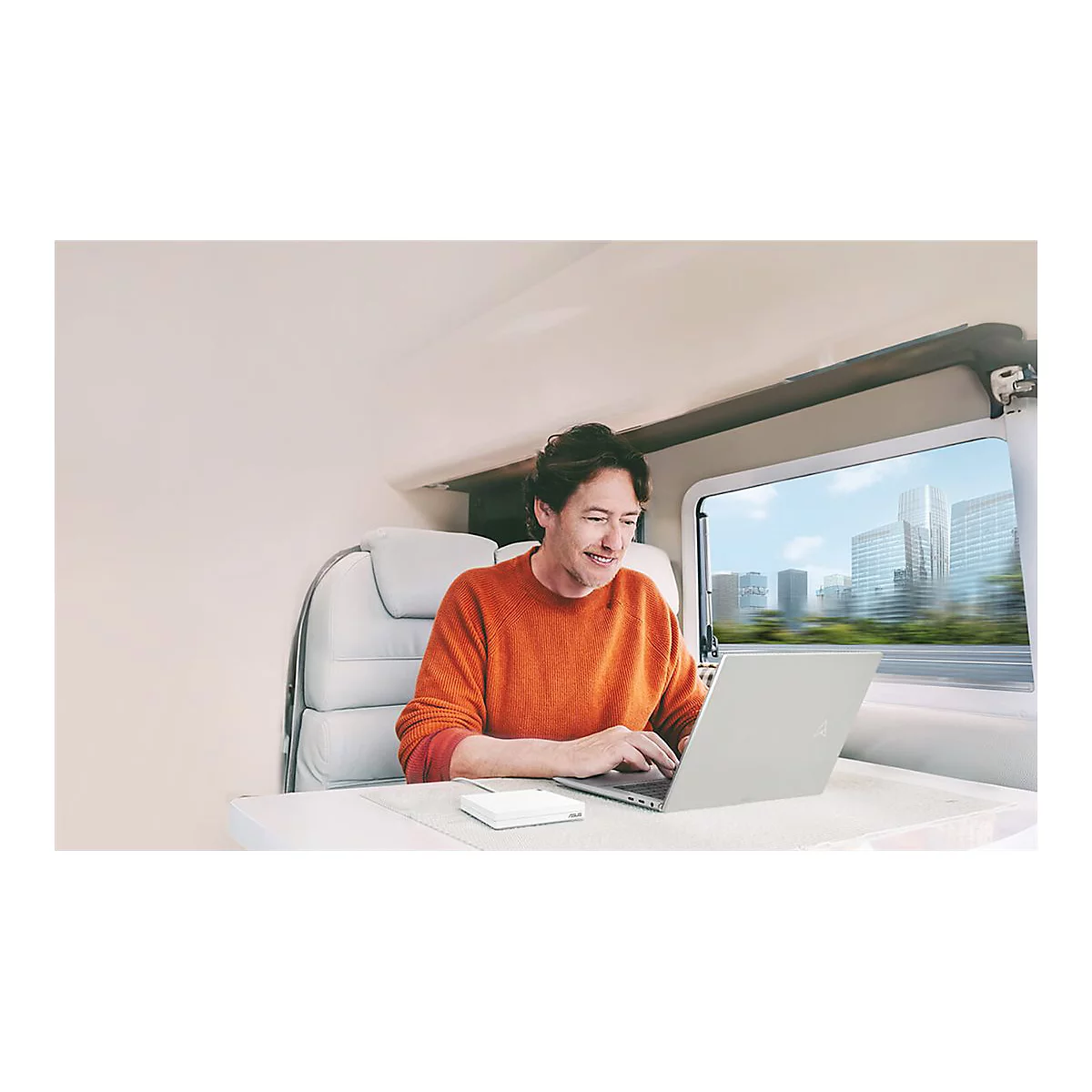 Ein Mann im Wohnwagen arbeitet mit Laptop und lächelt. Durch das Fenster ist eine Stadt zu sehen.