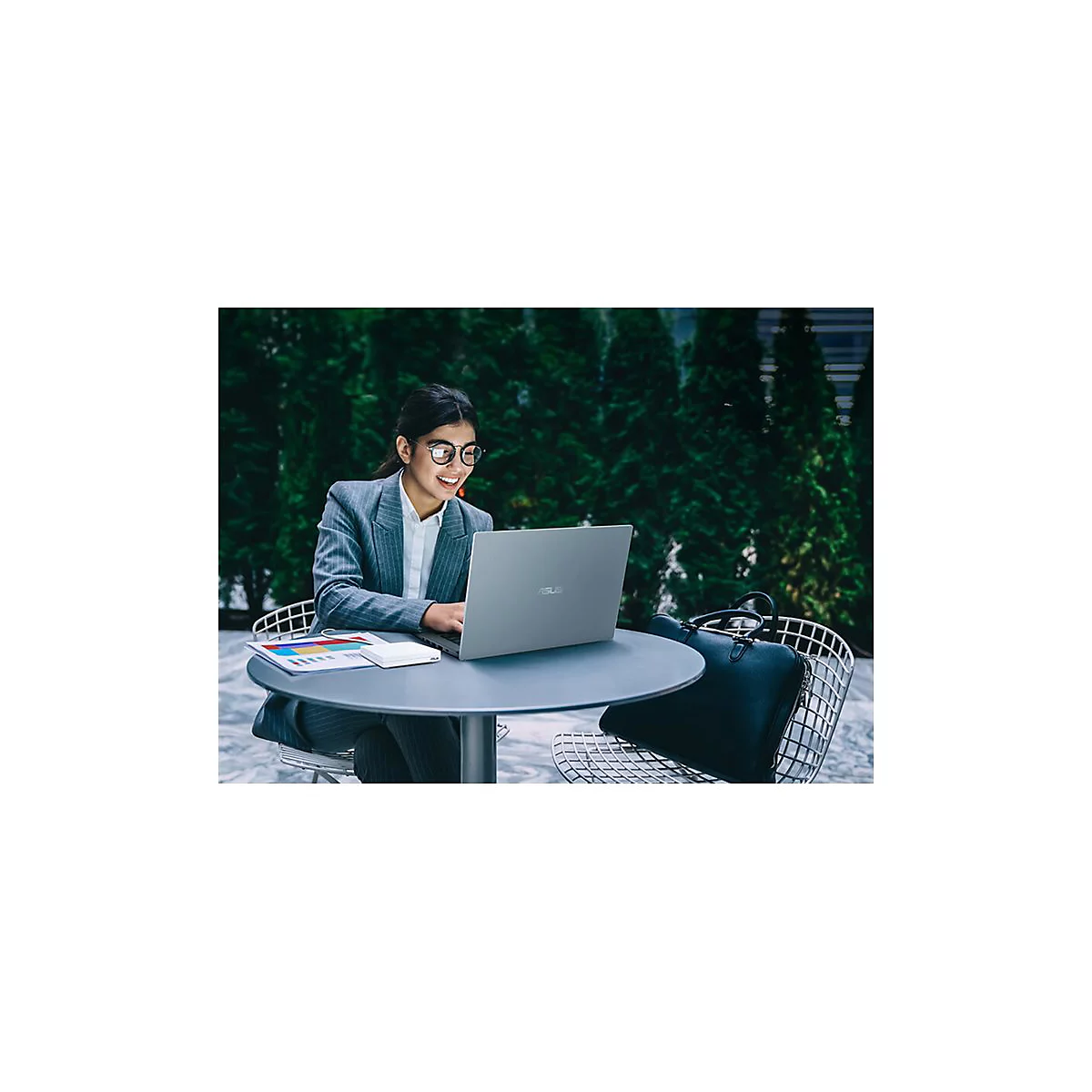 Junge Frau arbeitet mit Laptop an Tisch im Freien, lächelnd. Im Hintergrund Grünpflanzen und eine Aktentasche.