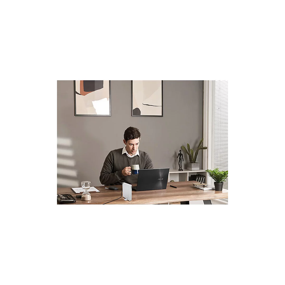 Ein Mann sitzt an einem Holzschreibtisch, Laptop, Tasse, weiße Box und Pflanzen.