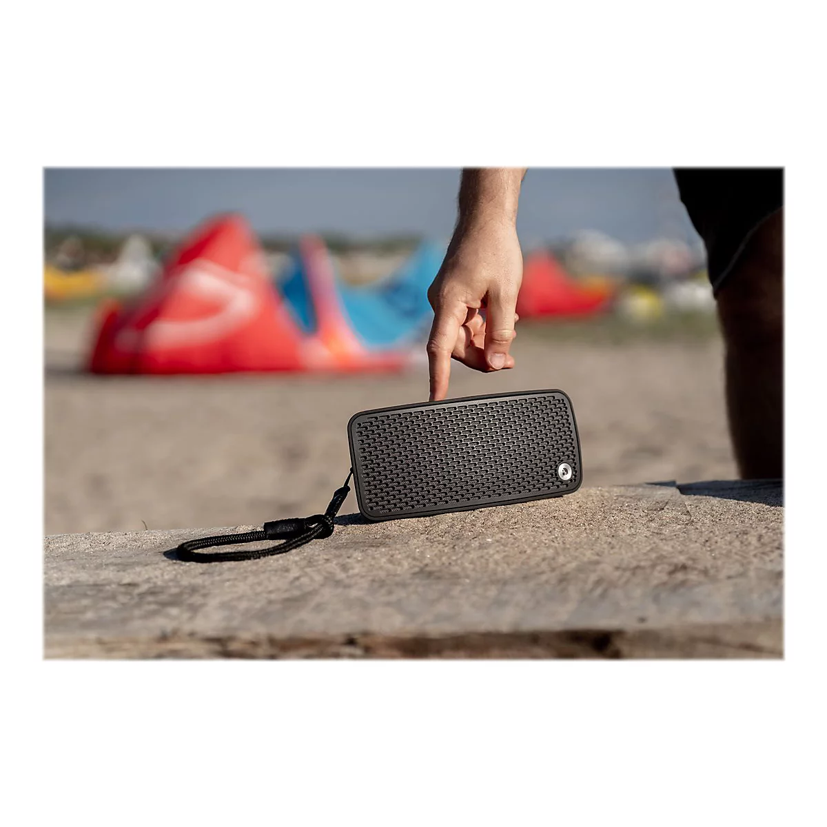 Ein Mann berührt mit dem Finger einen schwarzen Lautsprecher auf einem Stein. Im Hintergrund sind Strand und Kitesurfer.