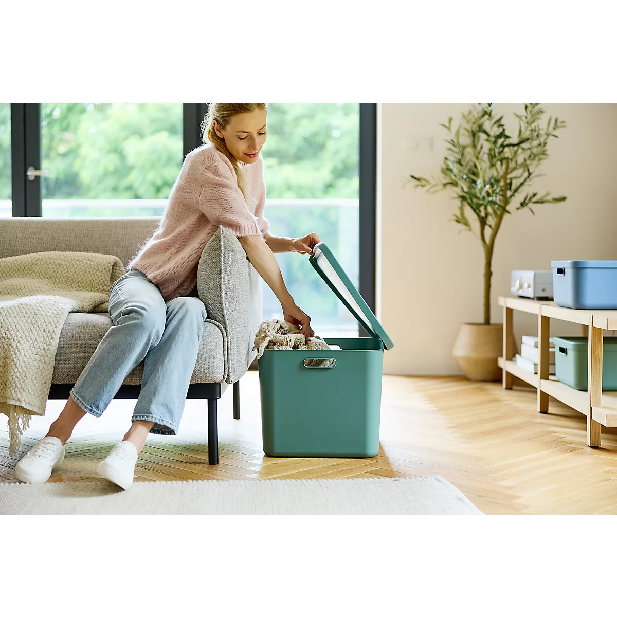 Türkisfarbene Aufbewahrungsbox steht neben Sofa, Holztisch und grosser Fensterfront.