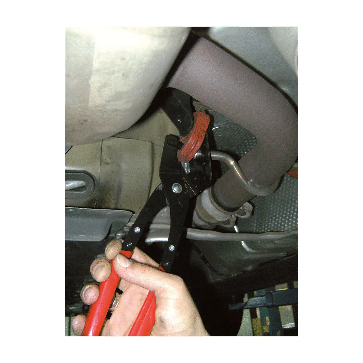 Nahaufnahme eines Autos von unten. Eine Person hält eine Zange mit roten Griffen, um eine Gummimanschette am Auspuff zu befestigen.