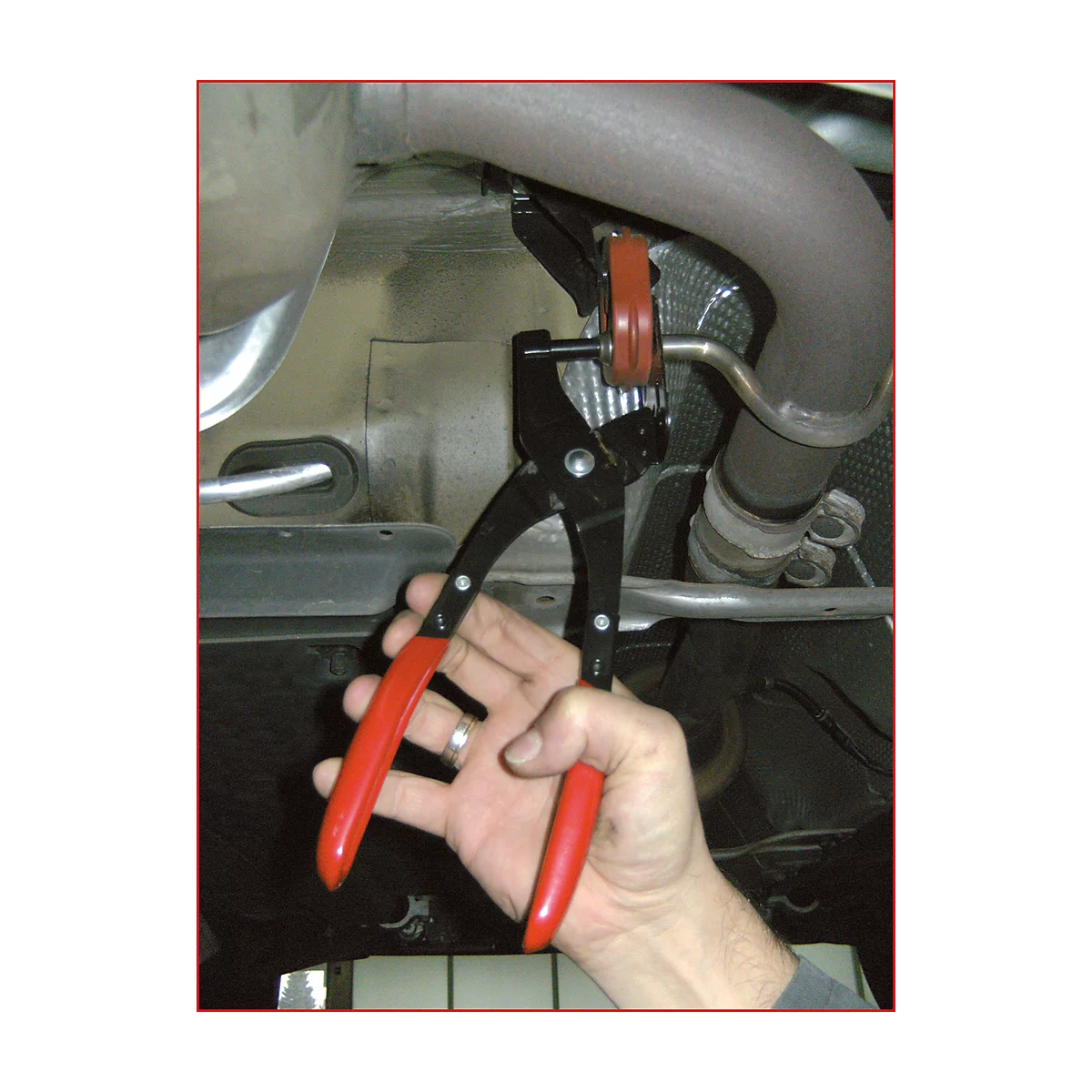 Hand hält Spezialzange am Auspuff. Schwarze Zange mit roten Griffen. Im Hintergrund Auspuff und Fahrzeugunterseite.