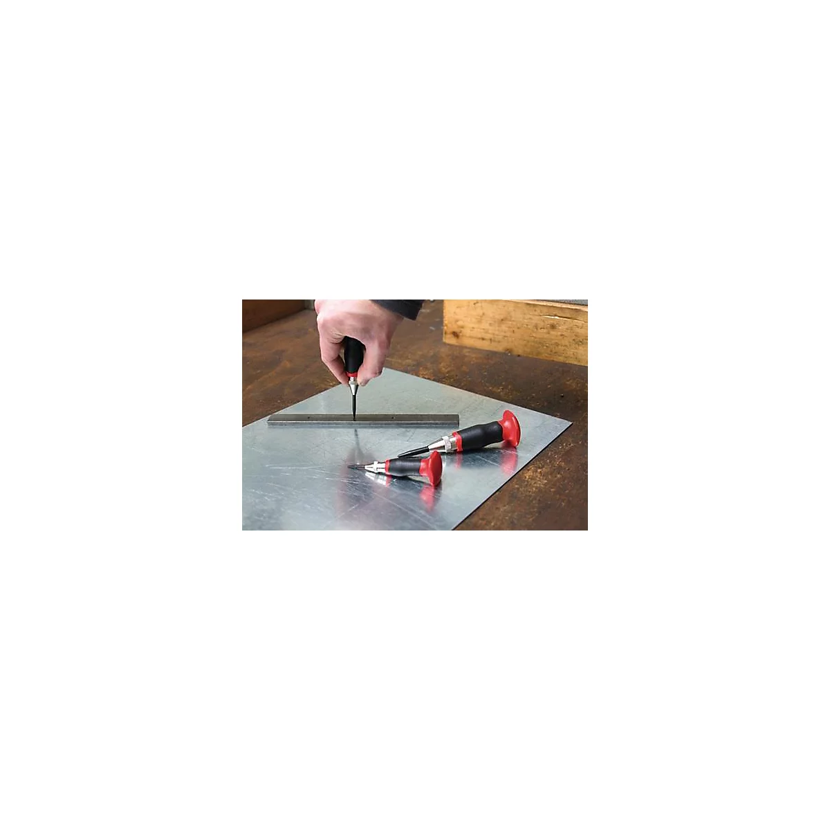 Een hand houdt een centerpons vast en zet een punt op een metalen plaat. Twee andere centerponsen liggen ernaast.