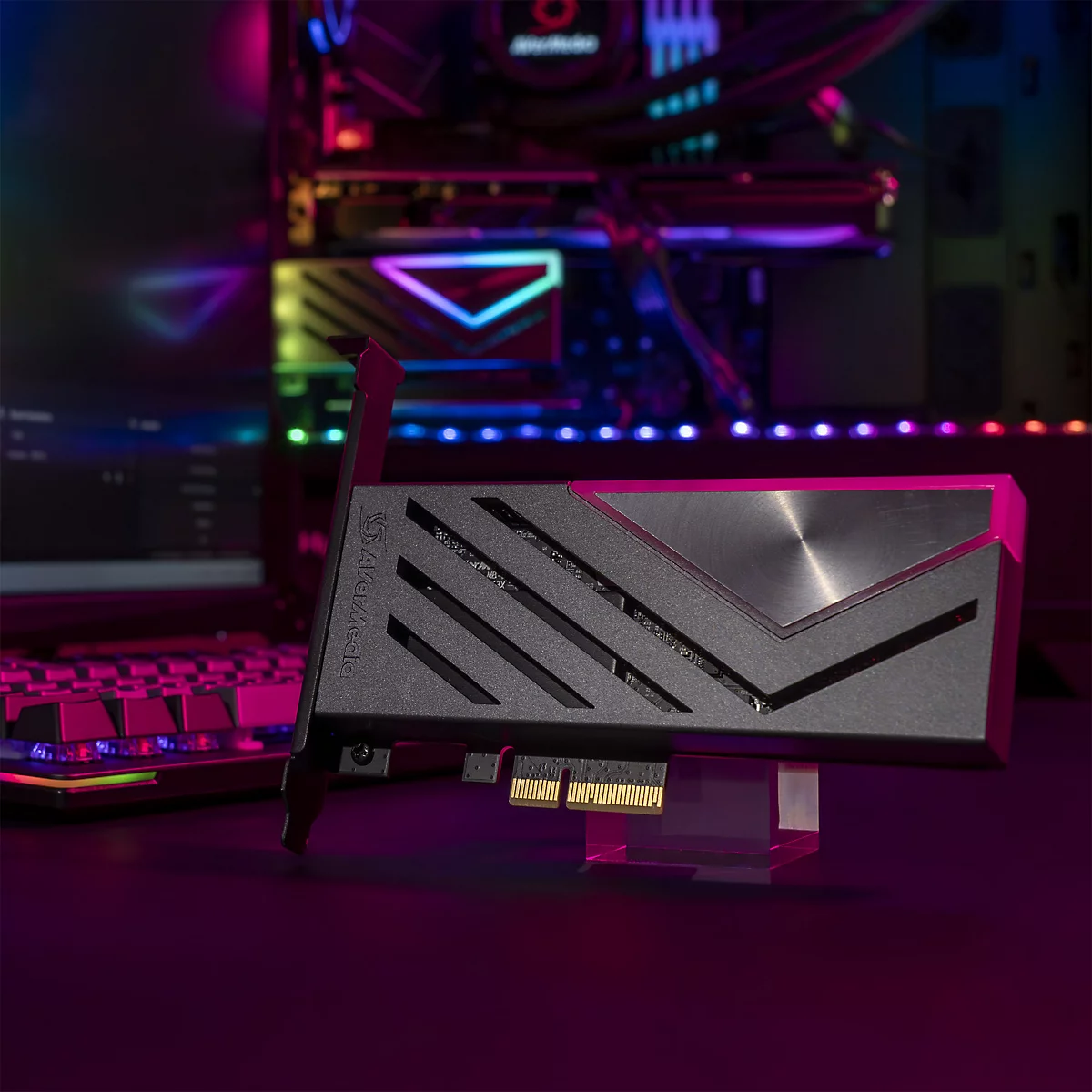 Schwarze AVerMedia PCIe-Capture-Karte vor einem Gaming-PC mit pinker Beleuchtung.