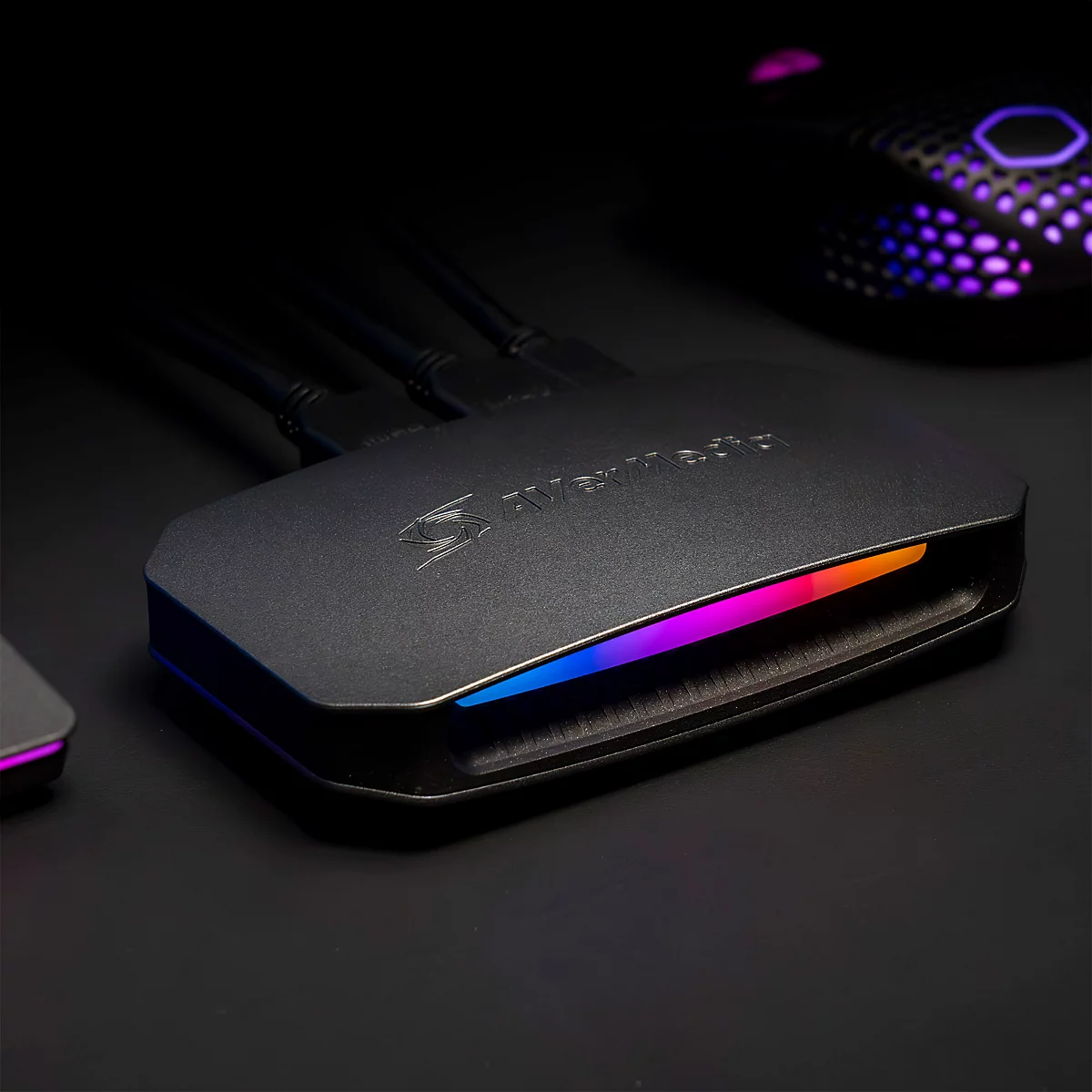 Schwarze AVerMedia Capture Card mit RGB-Beleuchtung auf einem dunklen Tisch, angeschlossene Kabel sichtbar.