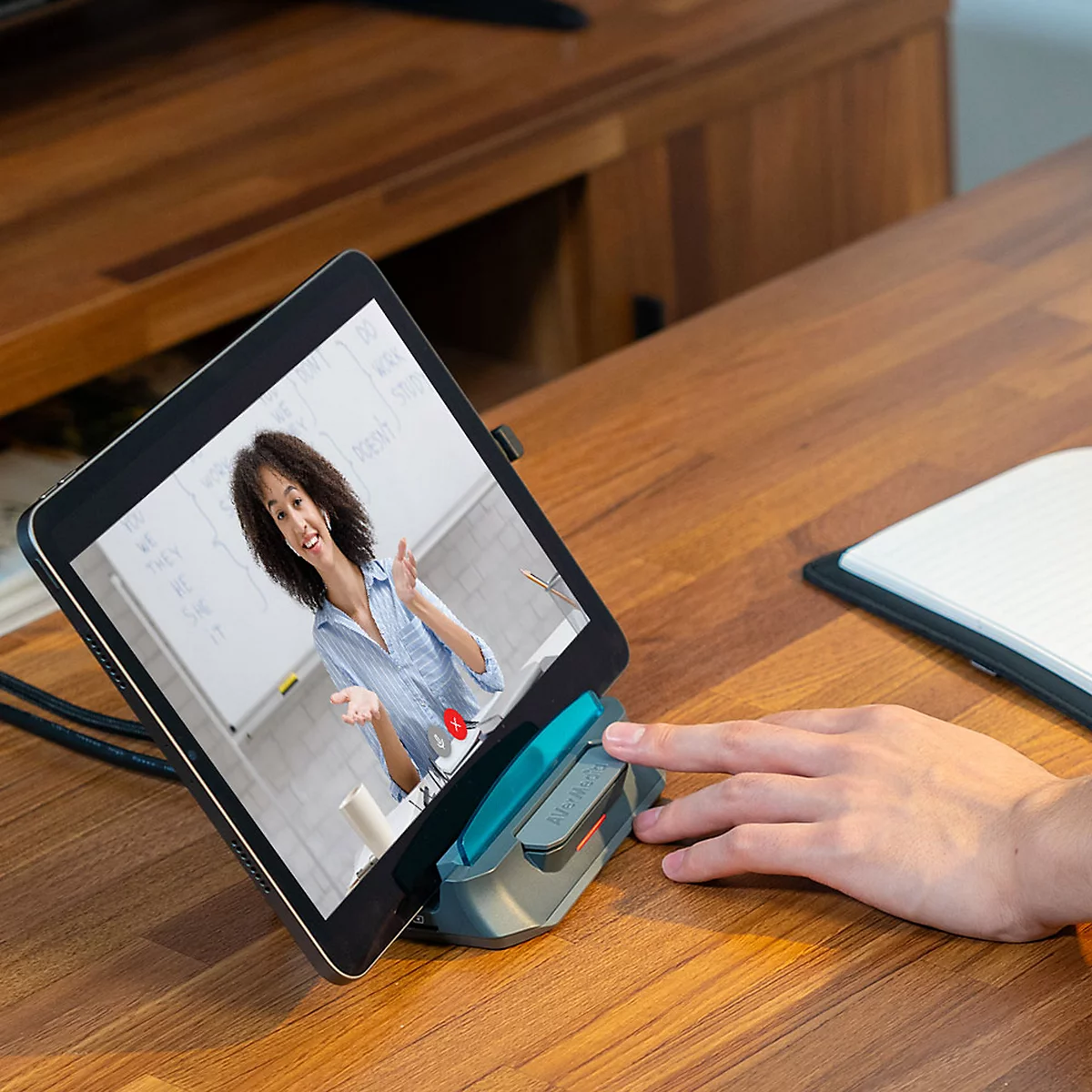 Ein Tablet in einem Standfuß auf einem Holztisch zeigt eine Frau bei einem Online-Unterricht per Videokonferenz.