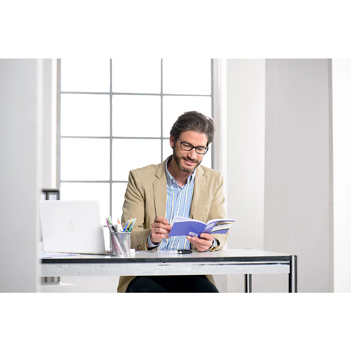A man with glasses reads a blue book at a desk in a bright office. He is wearing a beige jacket.
