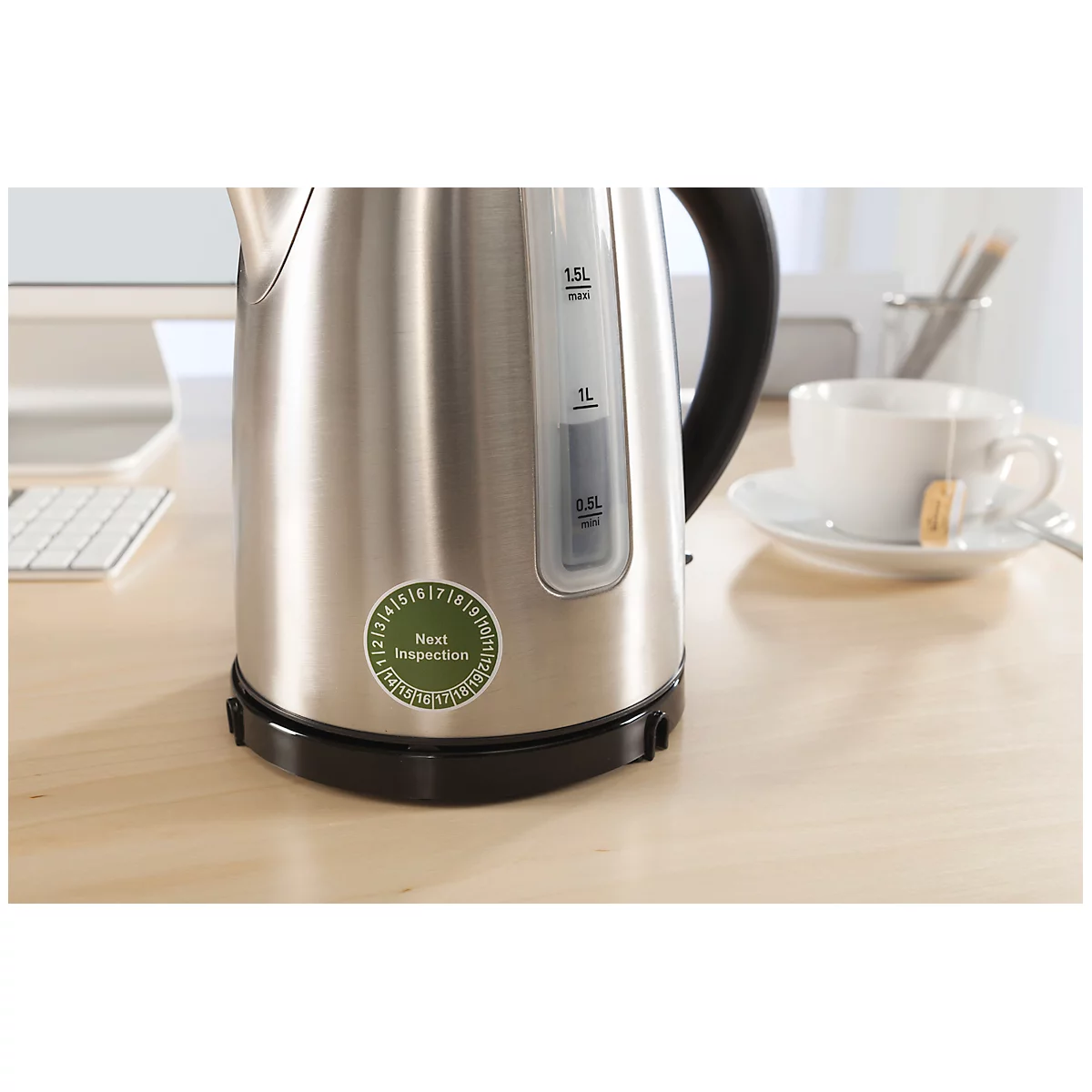 A stainless steel kettle sits on a table. Sticker "Next Inspection". A cup of tea next to it.