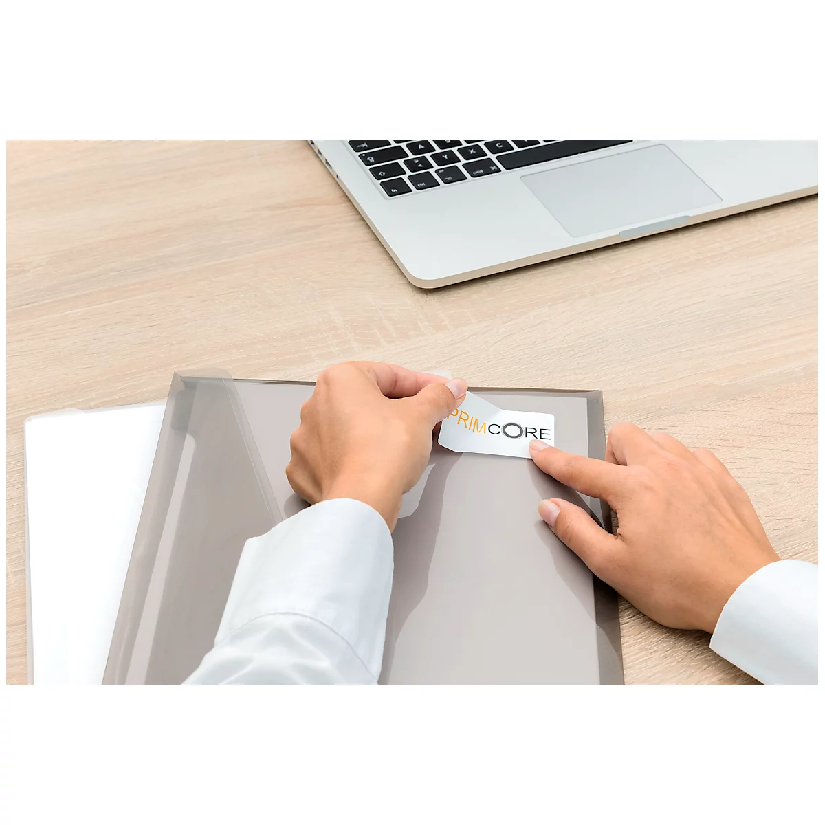 Person sticking a sticker with "PRIM CORE" on a folder. Laptop, folder and desk can be seen.