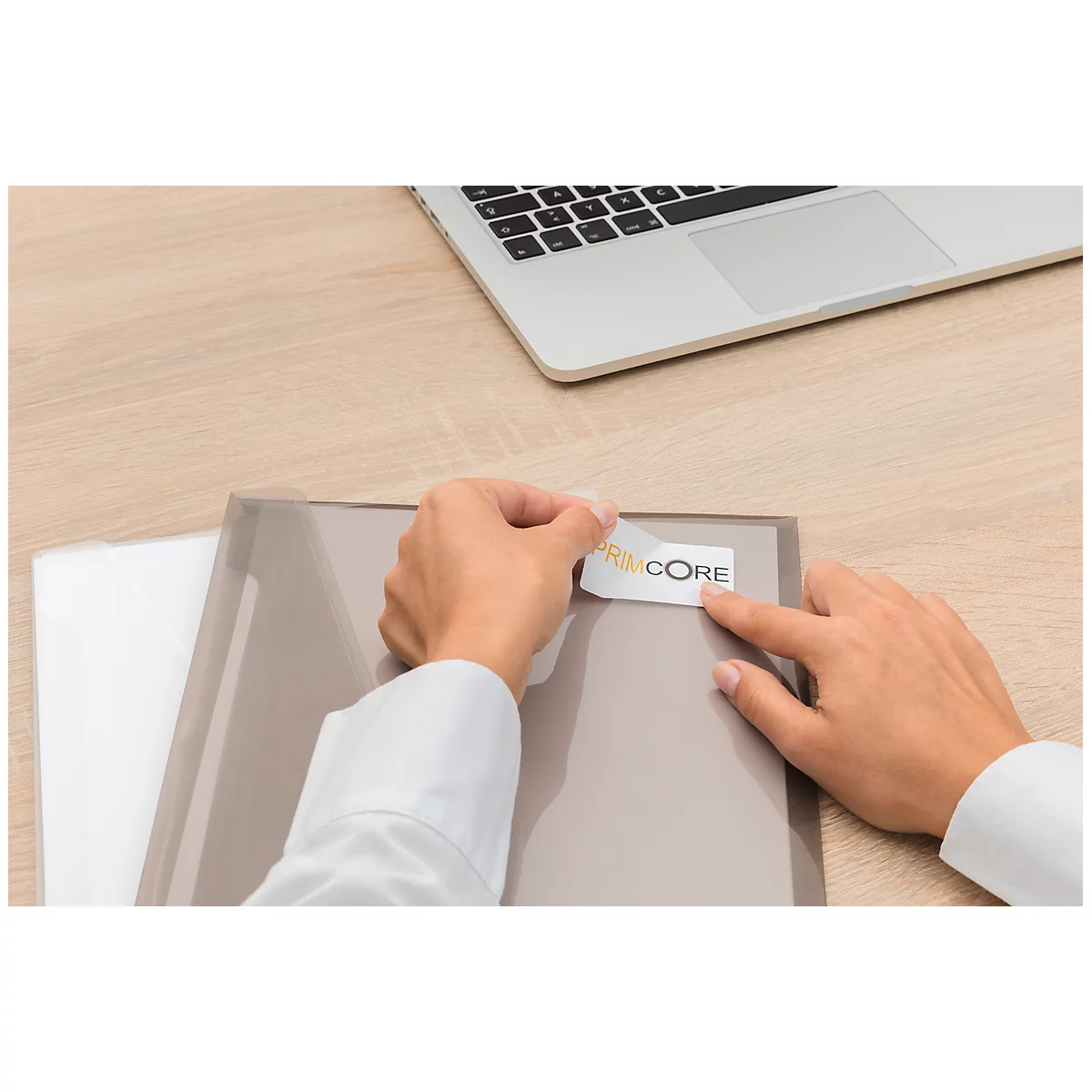Person sticking a sticker labeled "CORE" onto a gray folder. A laptop rests on the table.